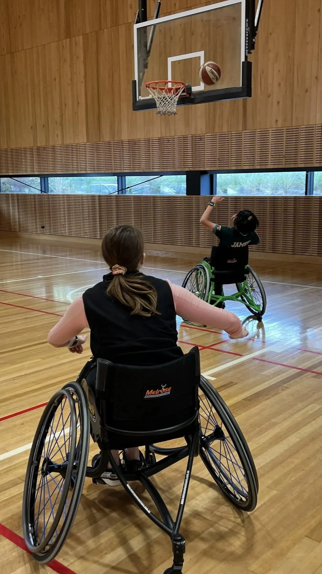 Two people in wheelchairs playing wheelchair basketball in a gymnasium. One person is shooting a basketball towards the hoop, and the other is watching.