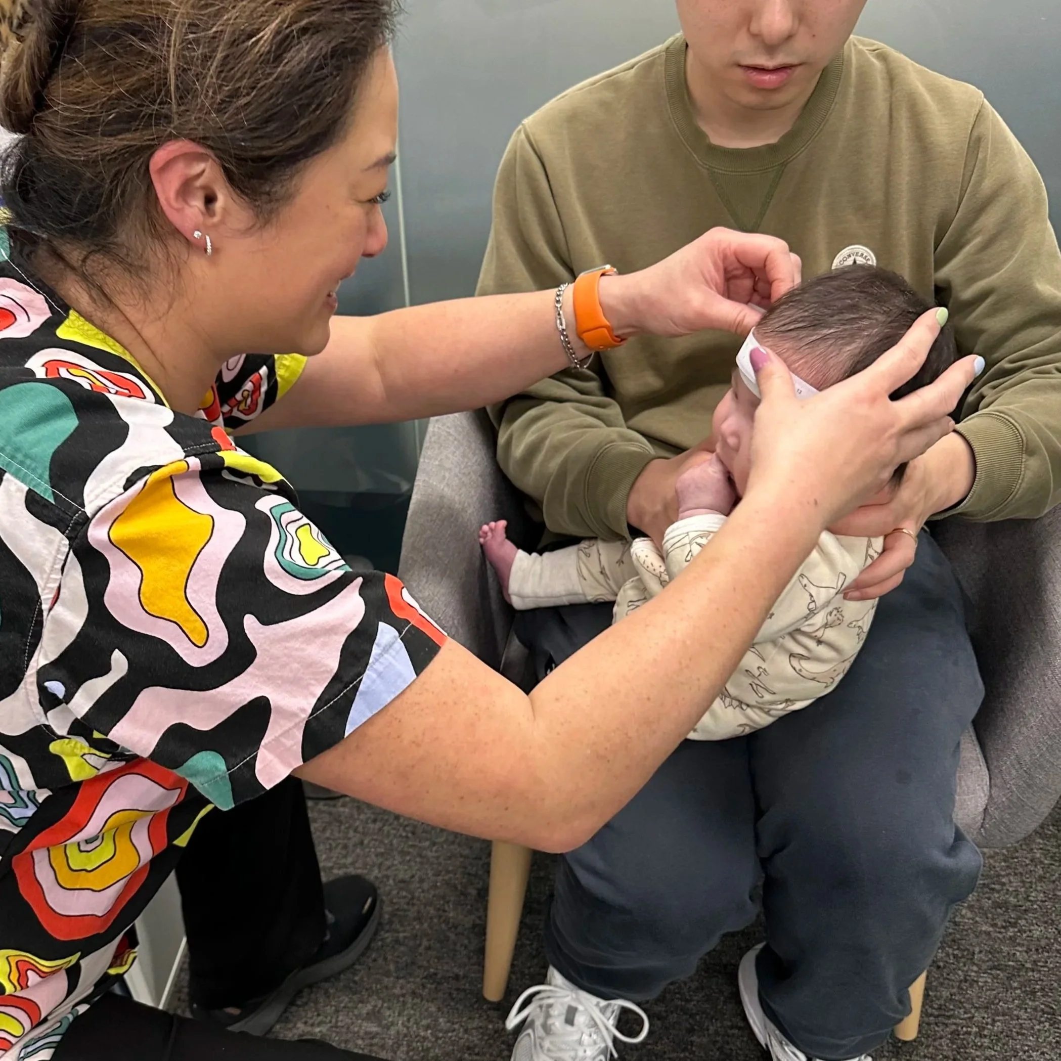 A nurse or doctor checks a baby's temperature with a forehead thermometer while the baby's father holds him on a gray armchair.