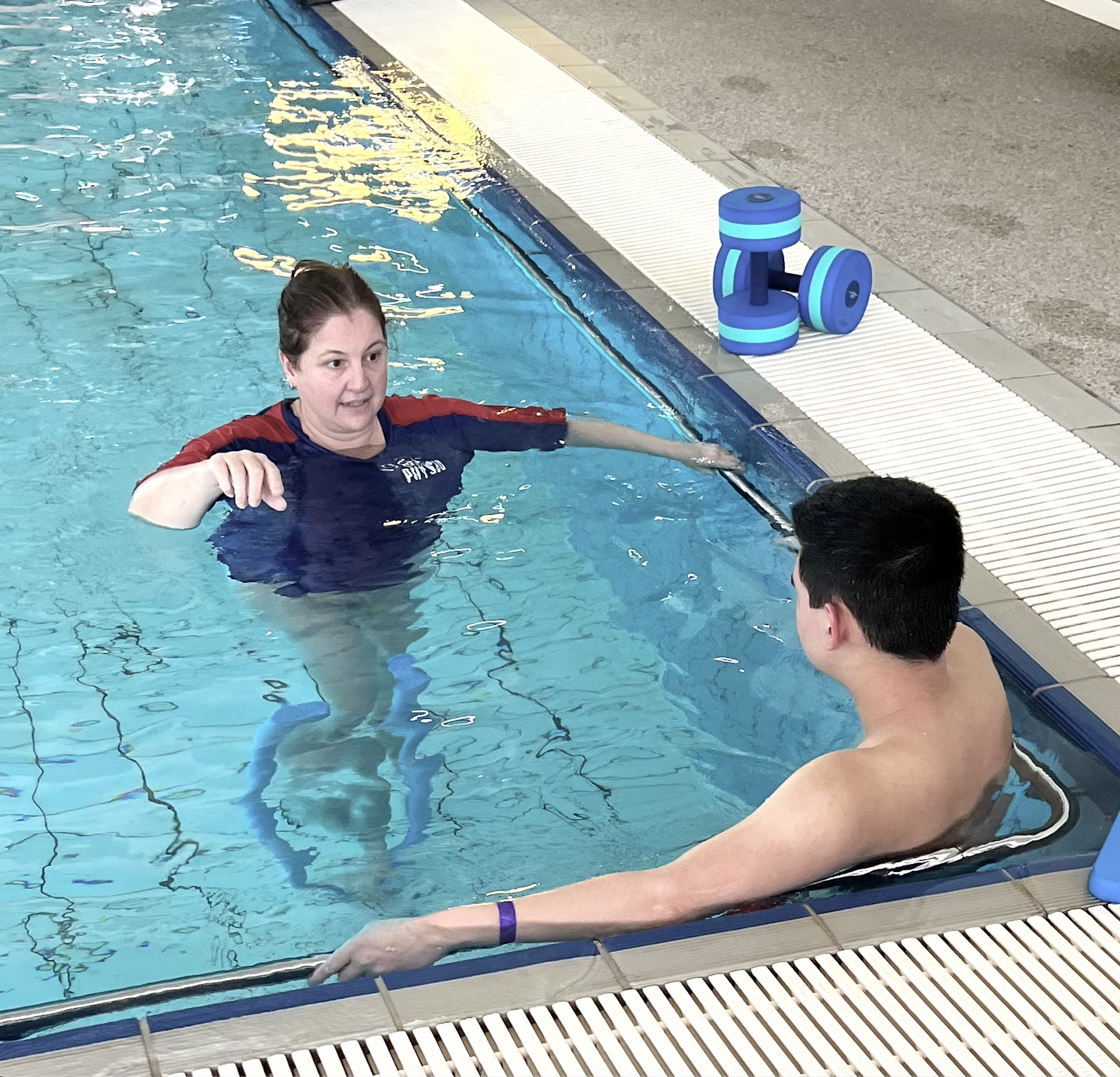 A woman in a blue and red shirt assisting a man in a swimming pool during a swim therapy session. Foam weights are placed on the pool deck.