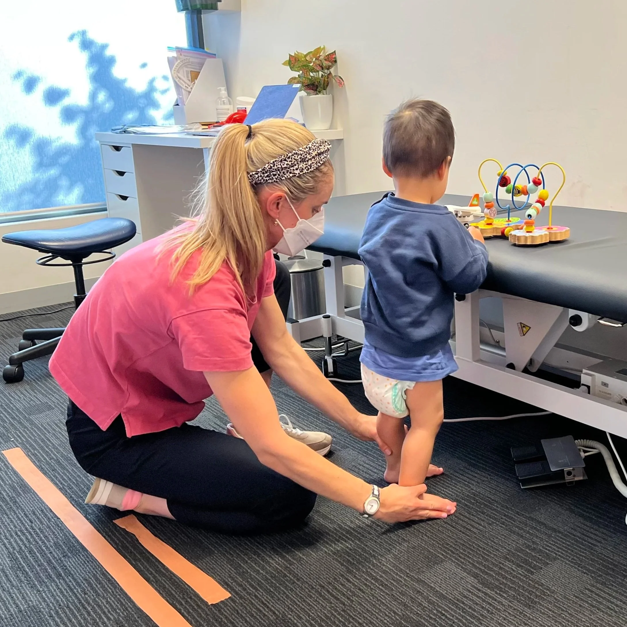A healthcare professional helping a young child walk in a medical examination room.
