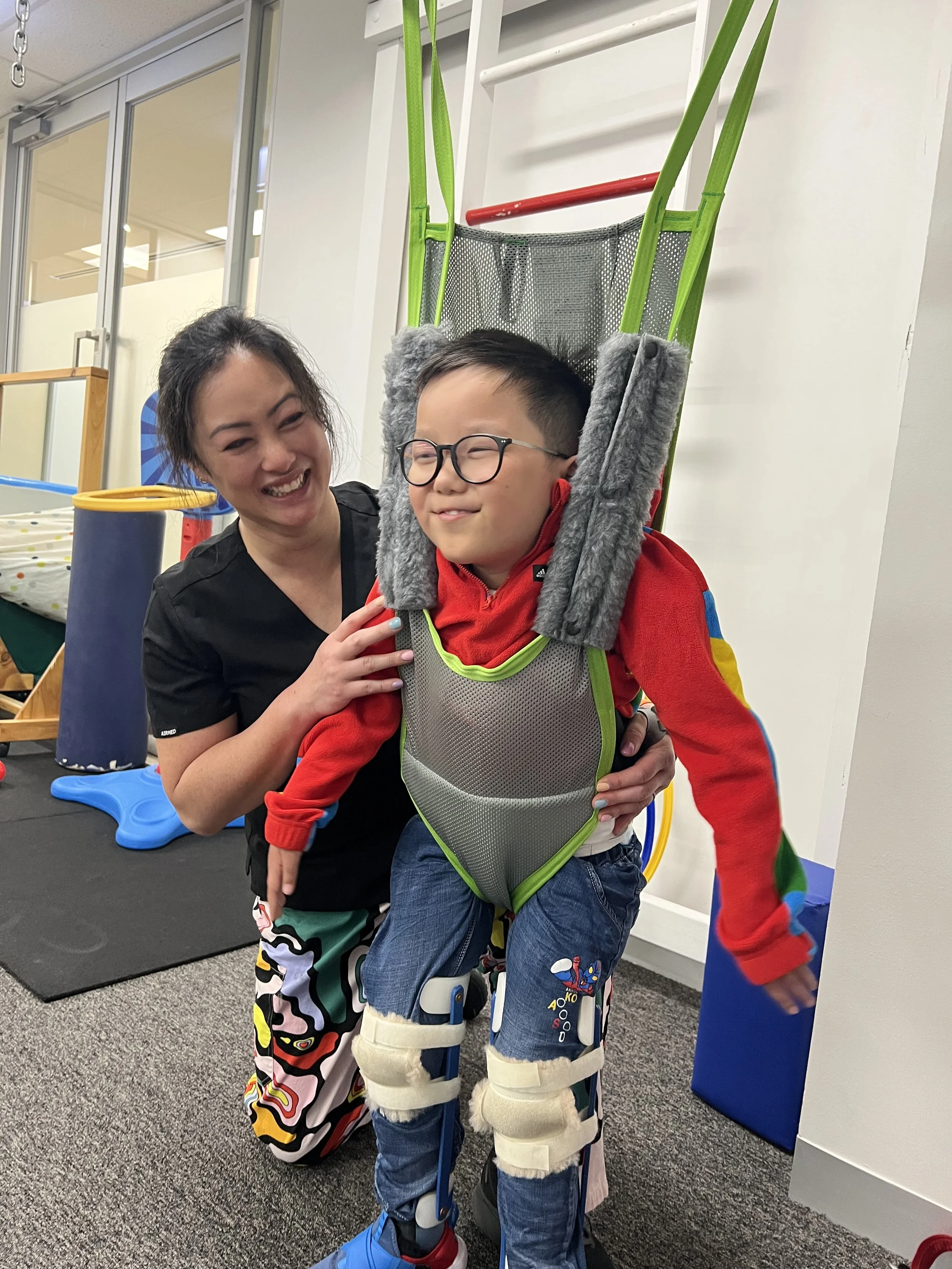 Child wearing leg braces in a supportive harness with a caregiver in a therapy or rehabilitation setting.