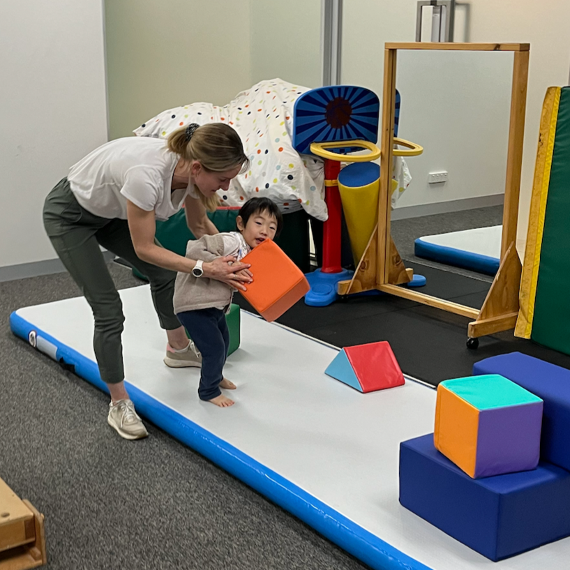 Child and adult playing on an indoor soft play mat with colorful blocks and gym equipment nearby.