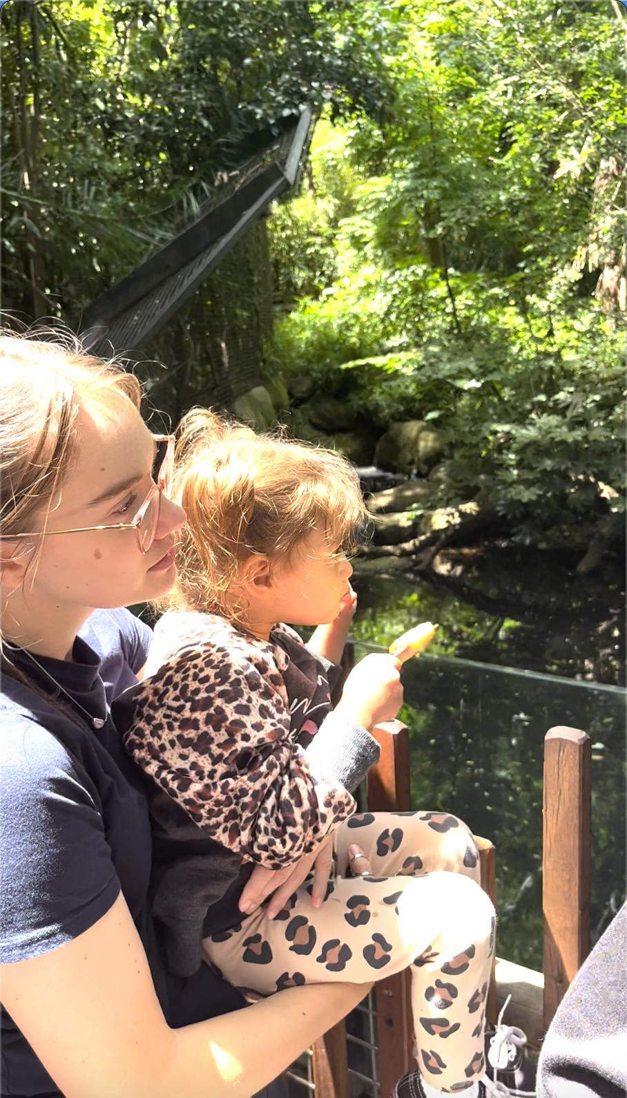 Two young girls sitting outdoors near a wooded area, one is holding a yellow popsicle, and both are looking towards the water below.