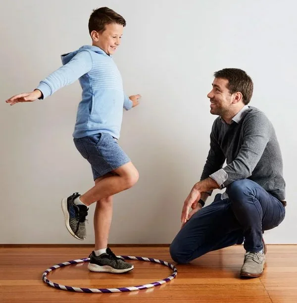 A boy jumping inside a hula hoop on the floor while an adult man kneels beside him, smiling, against a plain white wall.