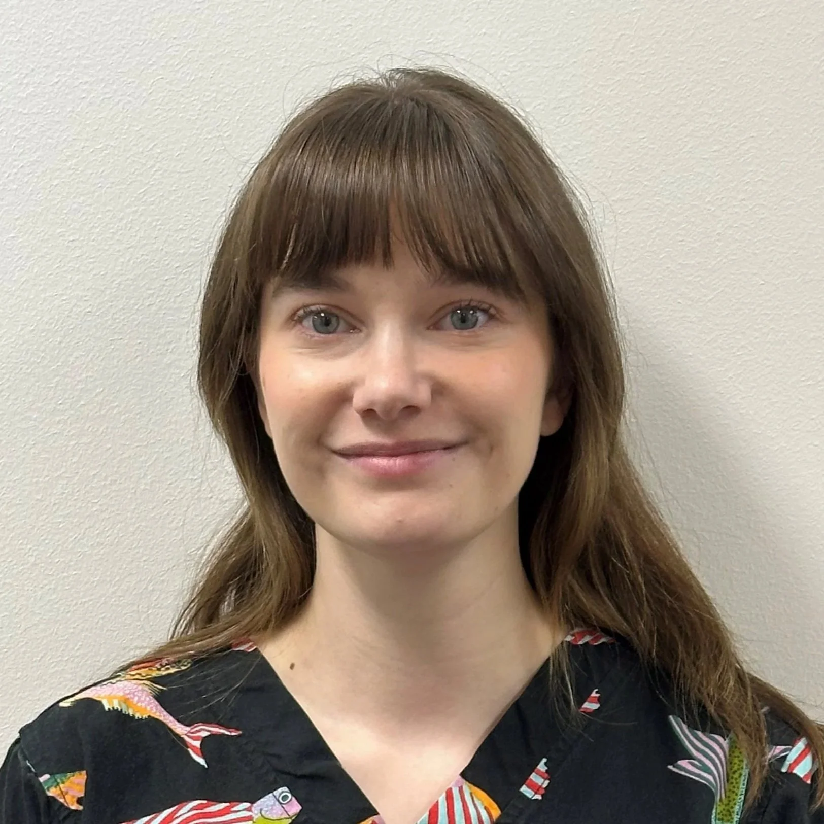 A woman with shoulder-length auburn hair, blue eyes, and light skin, smiling, standing against a plain beige wall, wearing a black top with colorful abstract illustrations.