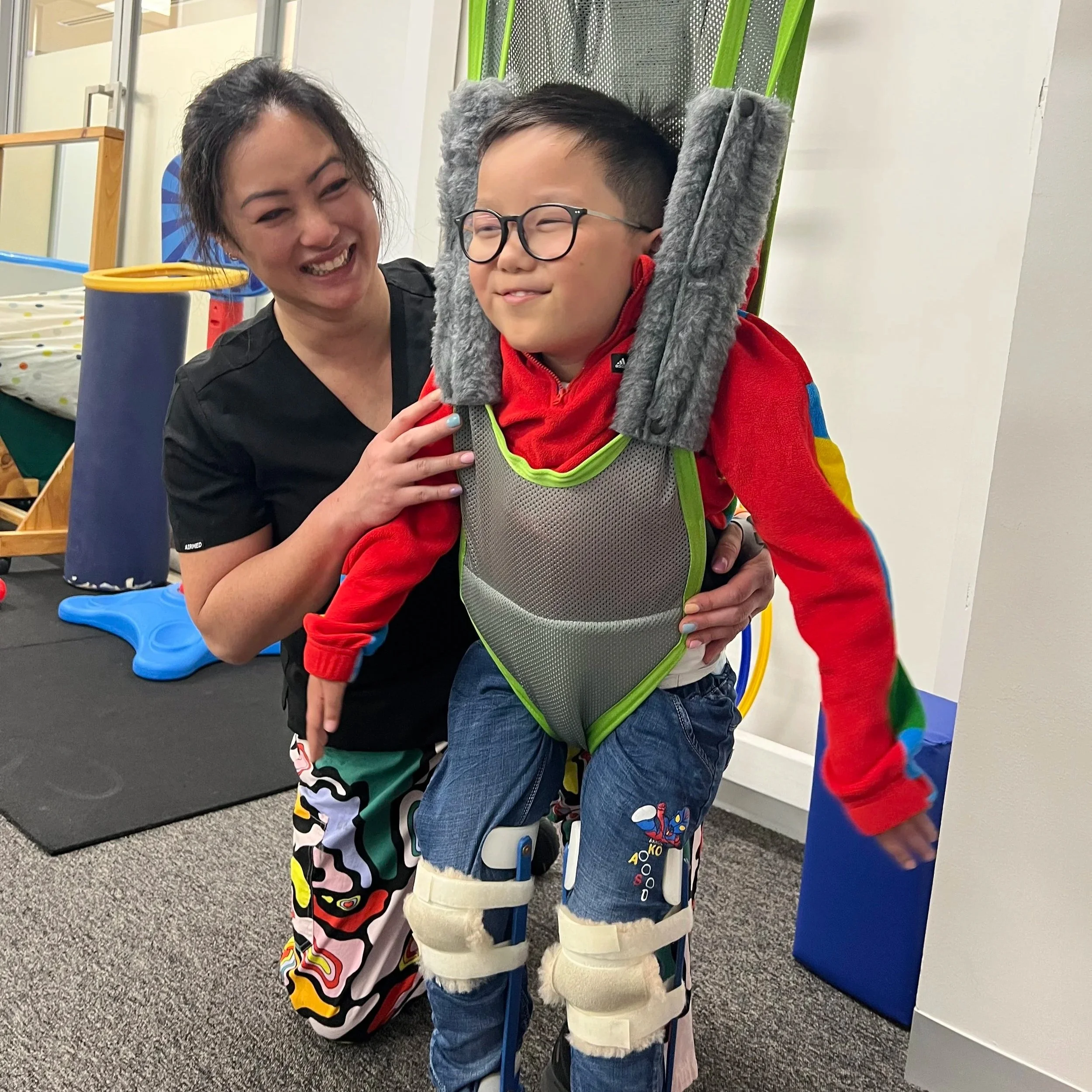 A smiling woman with curly hair and a young boy with glasses and a gray fur-lined harness, both indoors in a playful setting.