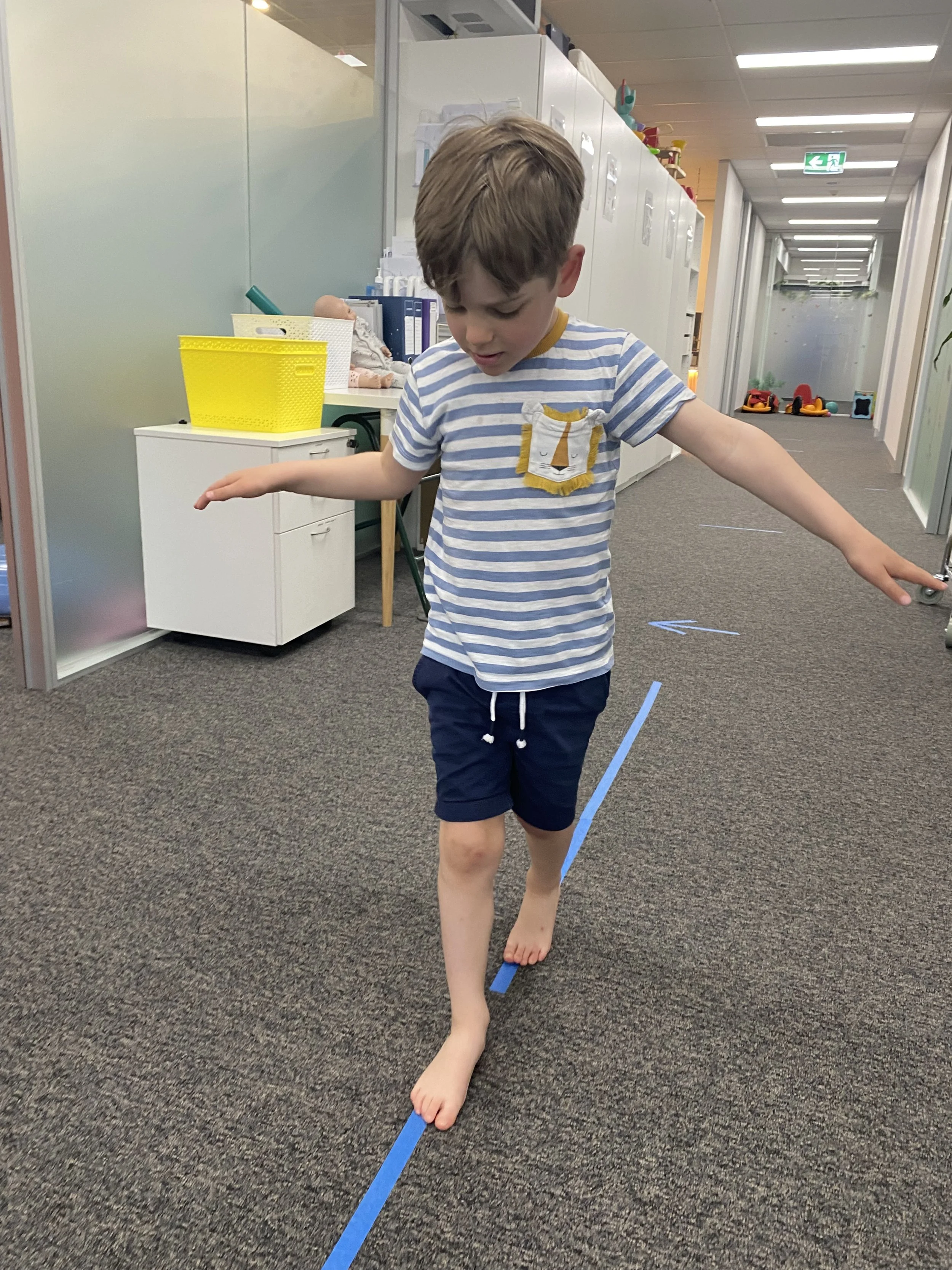 A young boy walking along a blue line taped on a gray carpeted floor in a hallway.