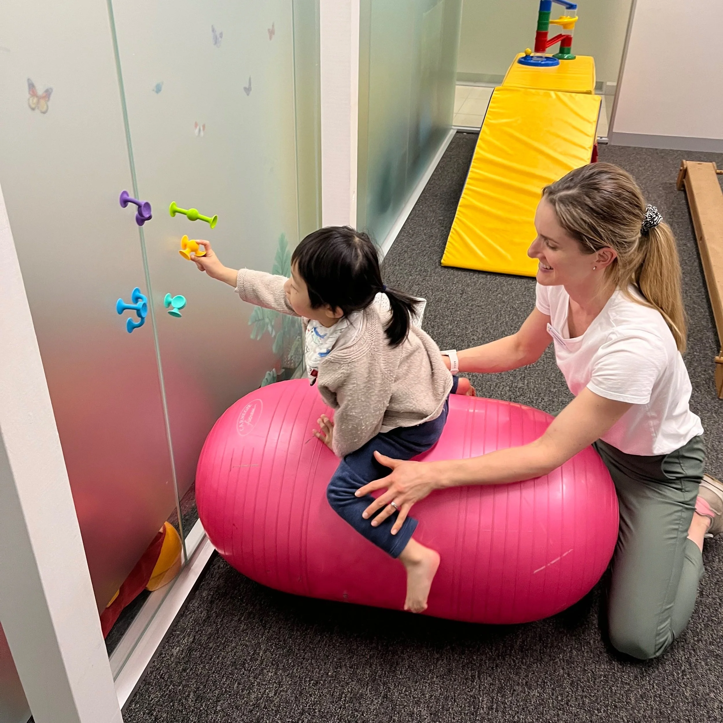 A woman helping a young girl practice standing on a pink exercise ball in a therapy or playroom, with colorful toys and wall decals of butterflies in the background.