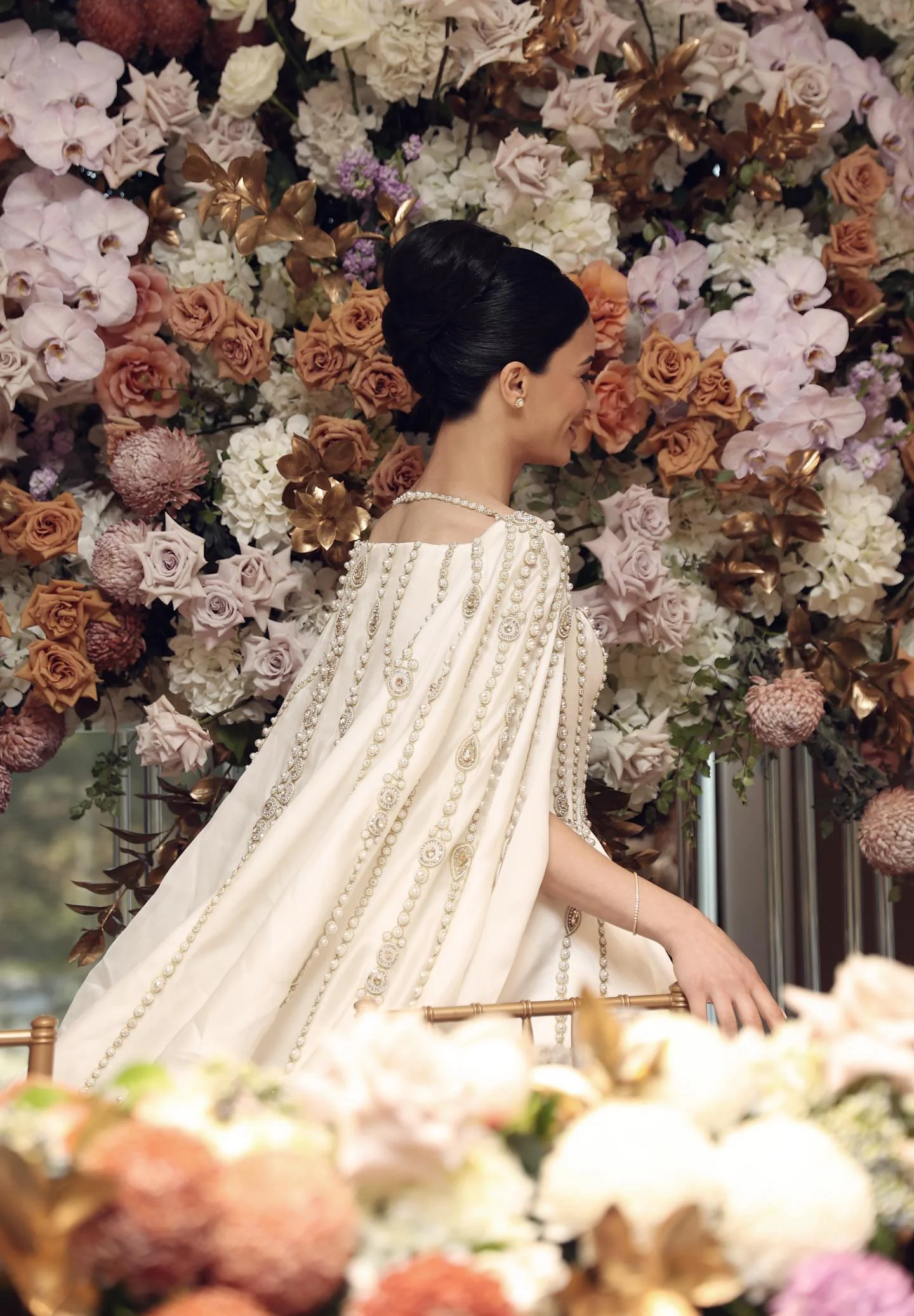 A woman with dark hair styled in an elegant updo, wearing a cream-colored dress with intricate pearl and embroidery details, and a pearl necklace and earrings, standing in front of a backdrop of colorful flowers, including roses, orchids, and greener