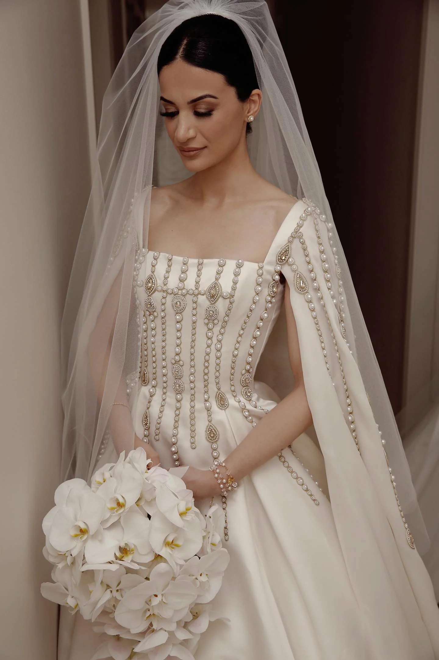 A bride with dark hair in an updo, wearing a pearl embellished wedding gown and veil, holding a bouquet of white orchids.