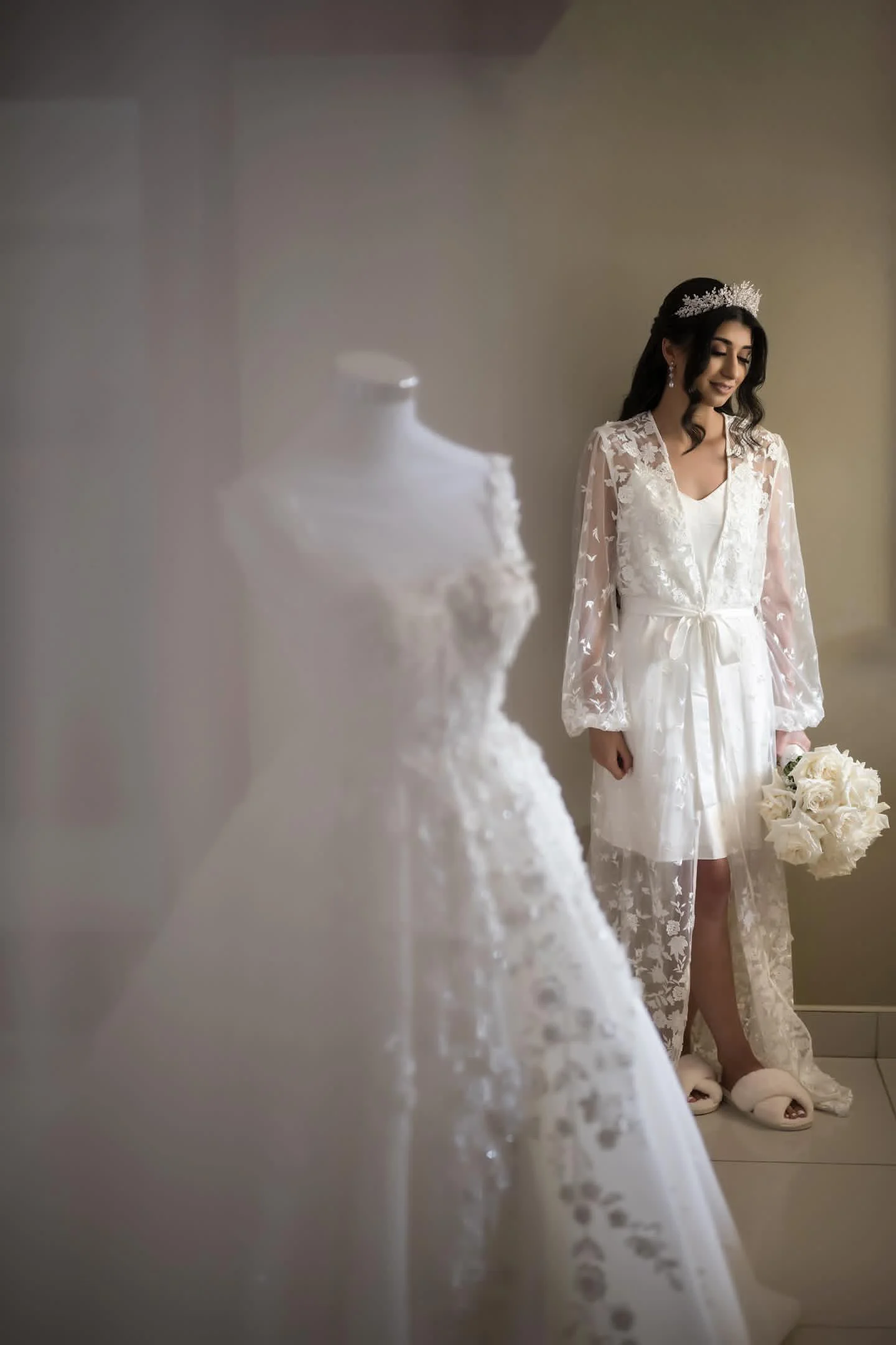 A bride in a white lace robe holding a bouquet of white roses, smiling with eyes closed, standing against a plain wall.