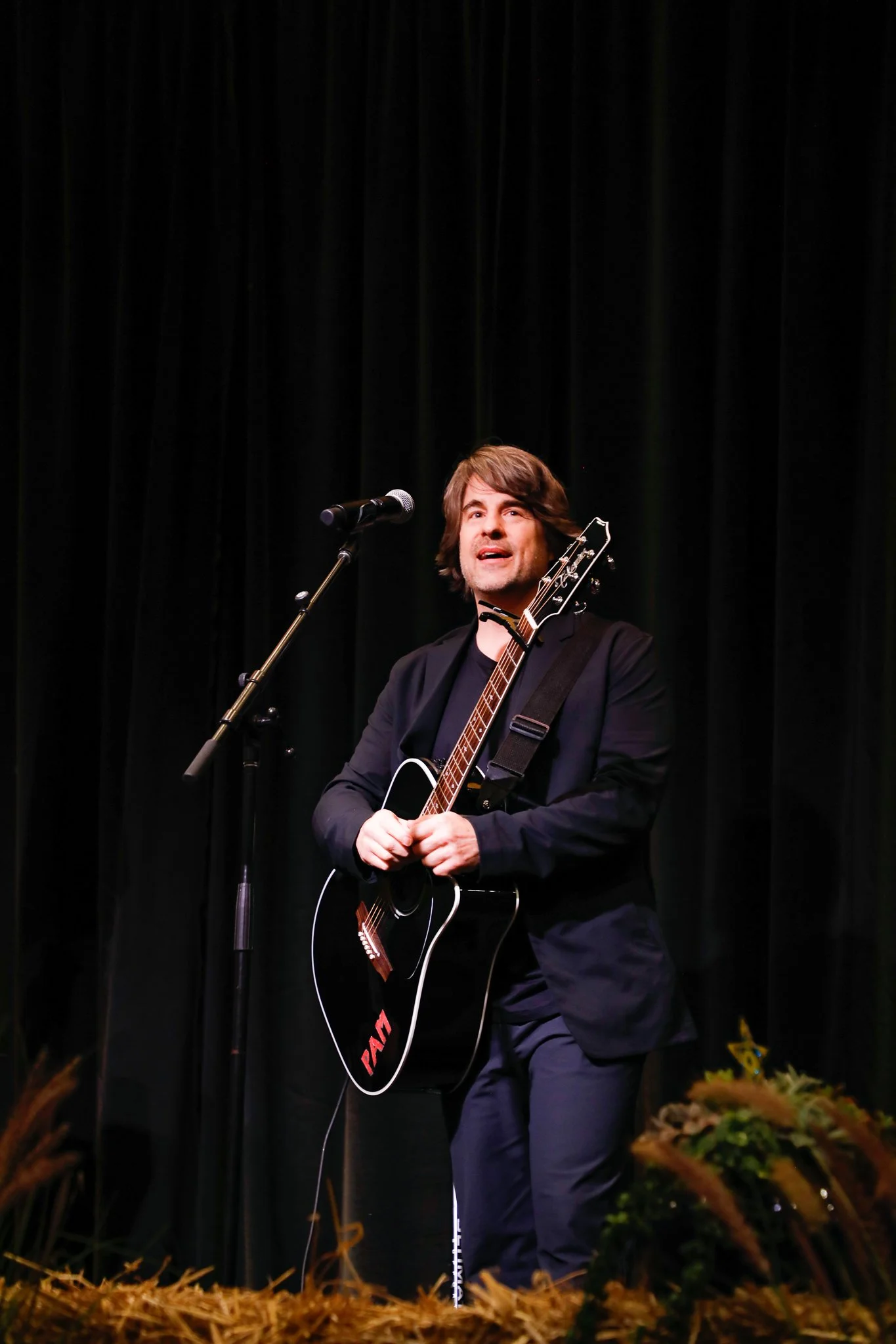 A male musician performing on stage with a guitar and a microphone in front of a black curtain.