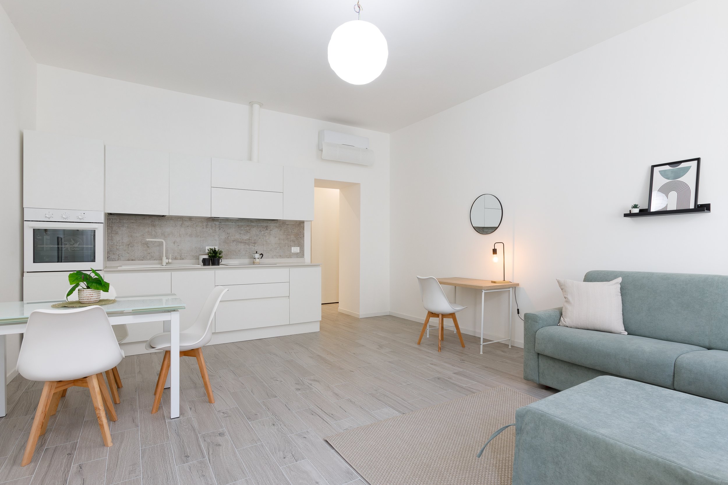 Interno di un appartamento moderno con cucina bianca, tavolo e sedie, divano grigio, piccolo angolo scrivania con lampada e specchio rotondo.