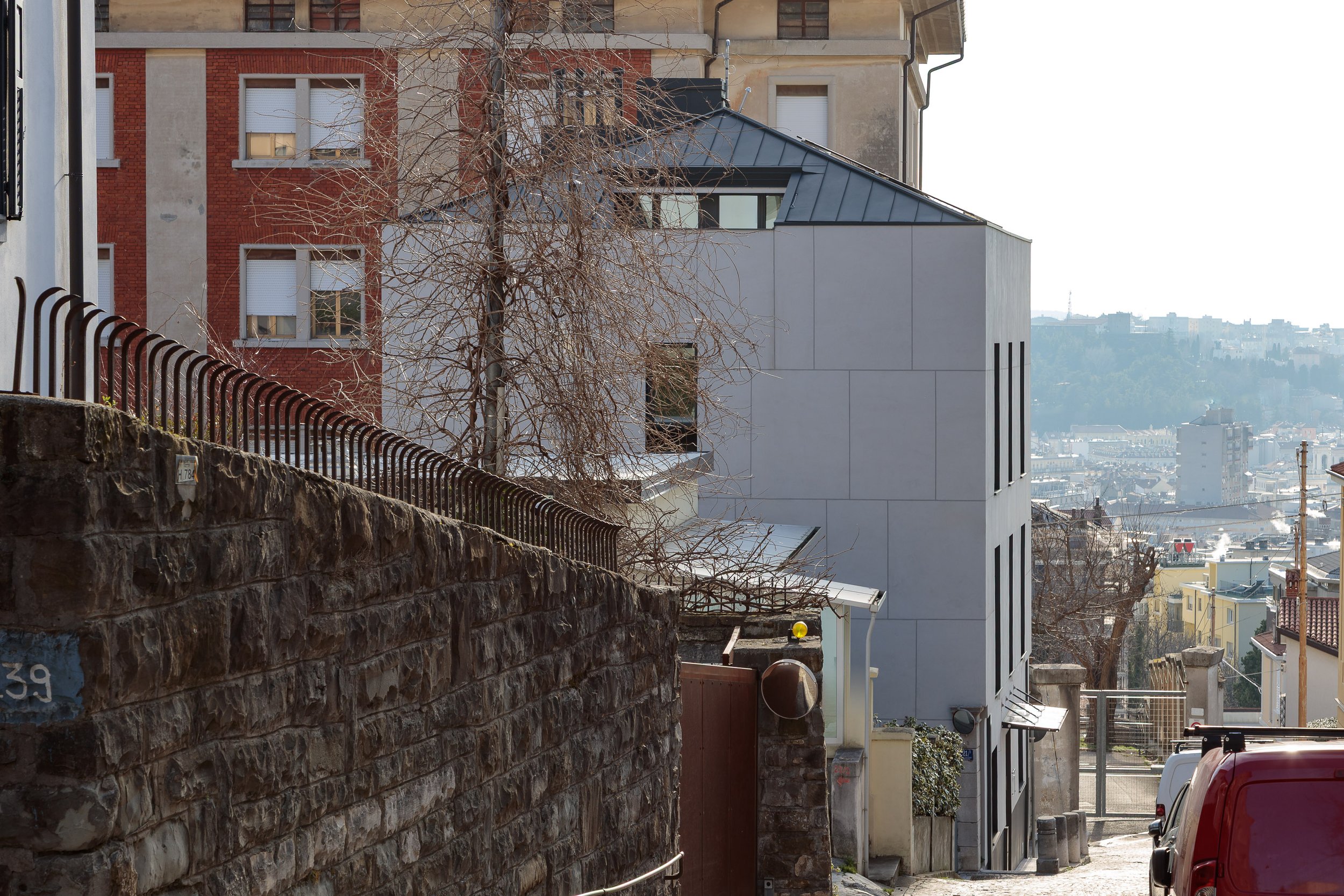 Vista di un quartiere urbano con edifici residenziali, un albero spoglio e ripida strada con muri di pietra e recinzioni in metallo, sullo sfondo si vedono altri edifici e colline.