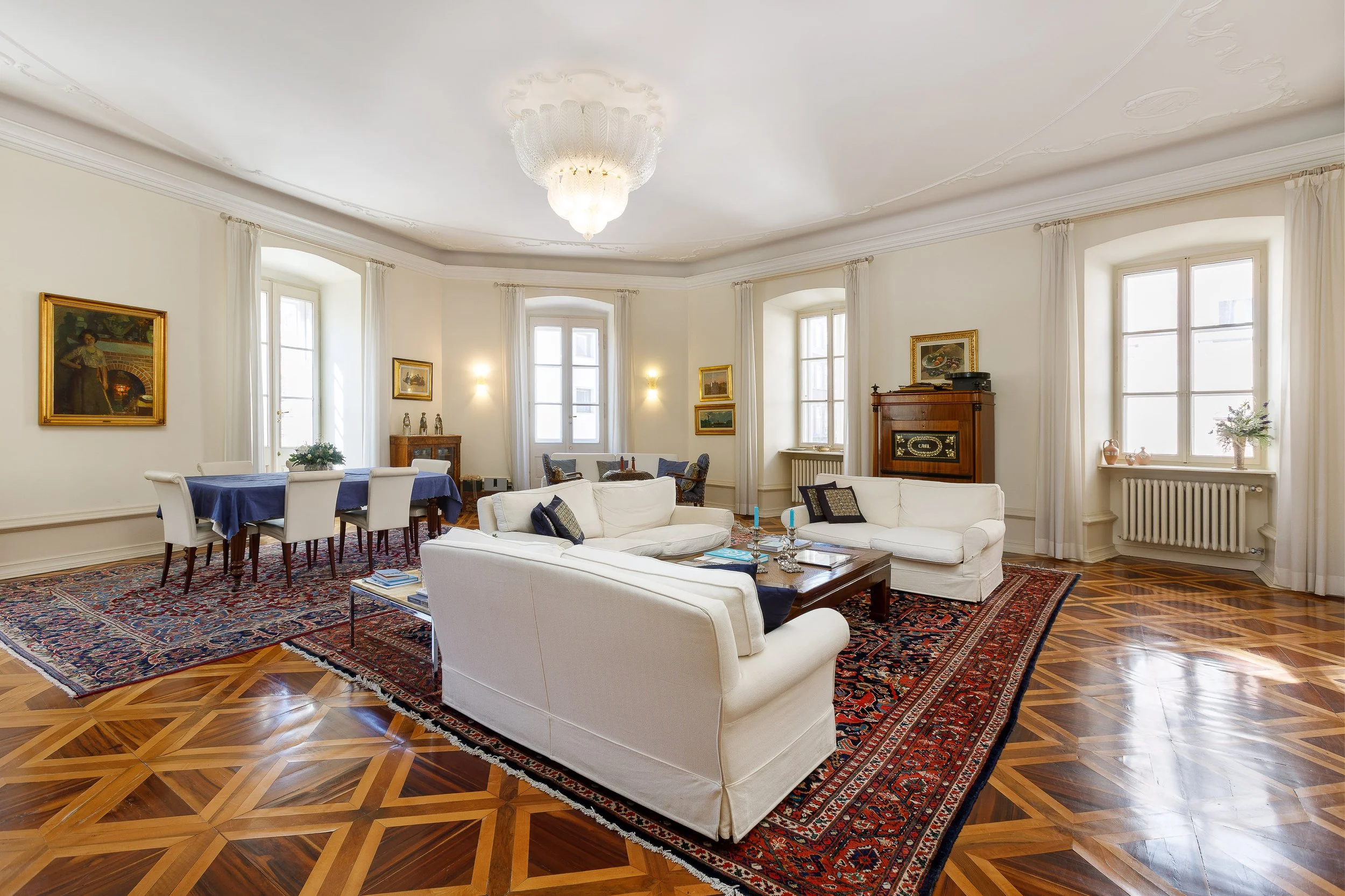 Salone elegante con pareti bianche, grandi finestre, tappeto orientale, divani bianchi, tavolo con tovaglia blu, quadri alle pareti, mobili in legno, lampadario chandelier di cristallo, decorazioni e piante.