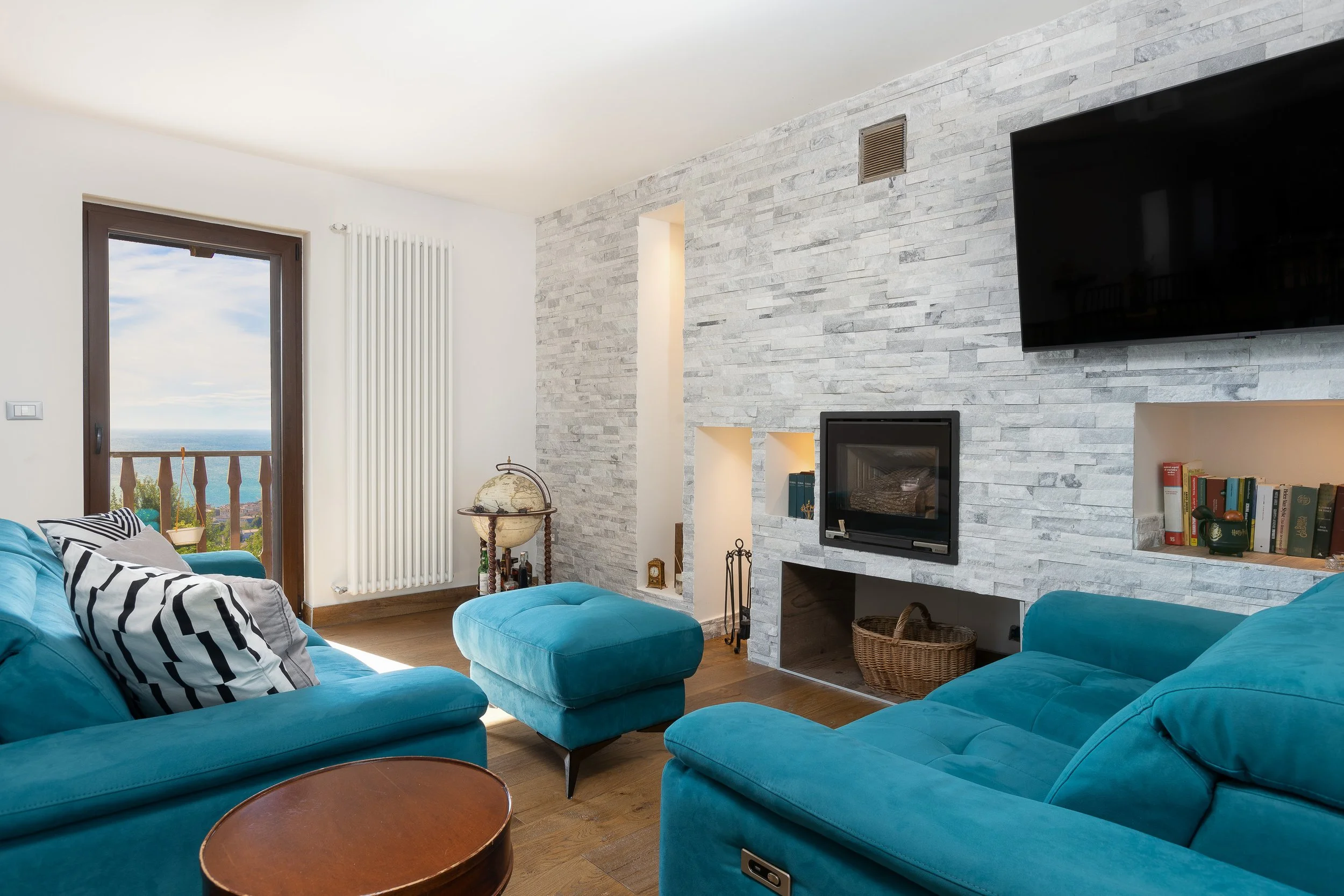 Interno soggiorno con divani blu, camino, libreria e TV, finestra con vista sul mare.