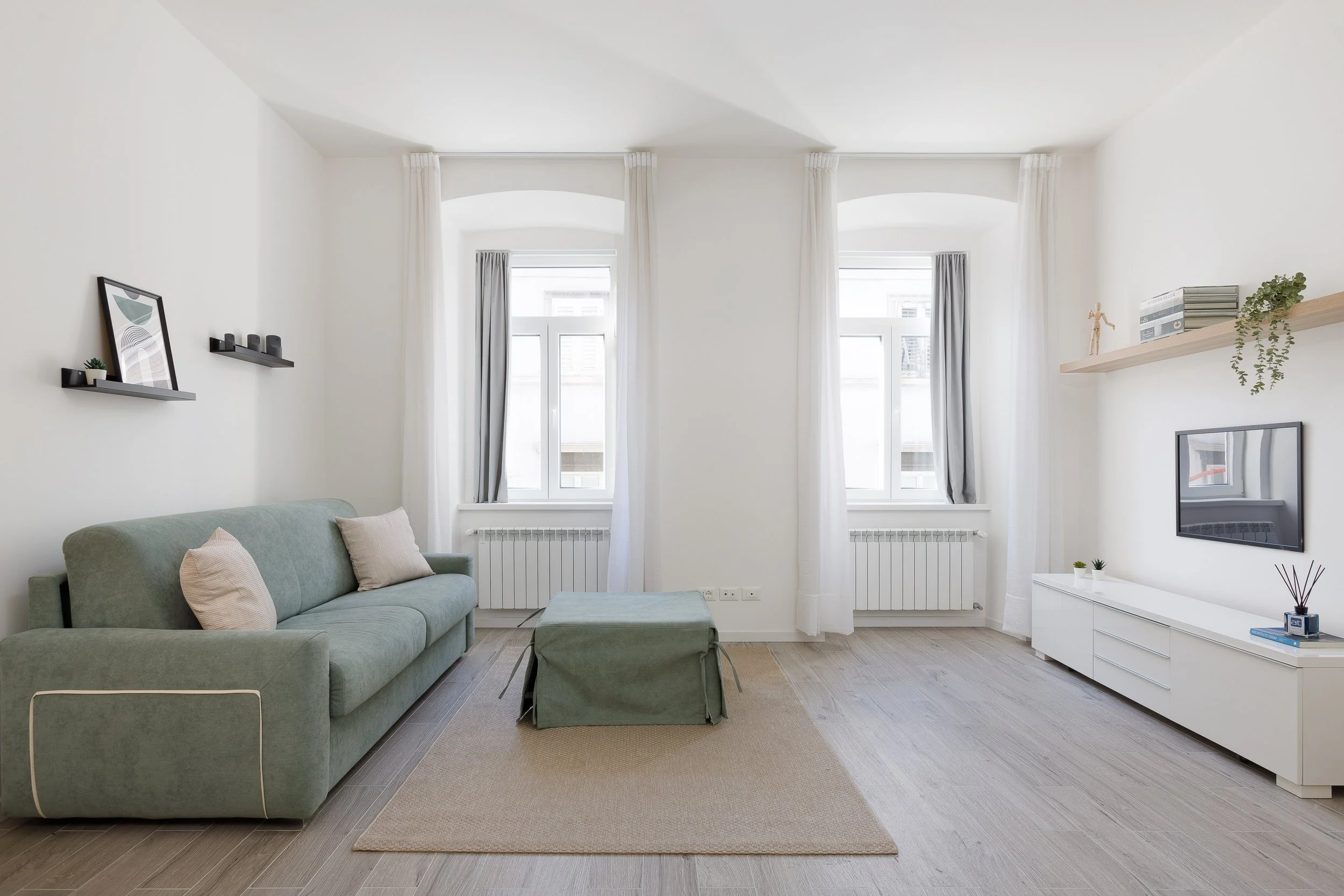 Soggiorno moderno con un divano verde, due finestre con tende bianche, un mobile TV bianco, mensole con decorazioni e pavimento in legno chiaro.