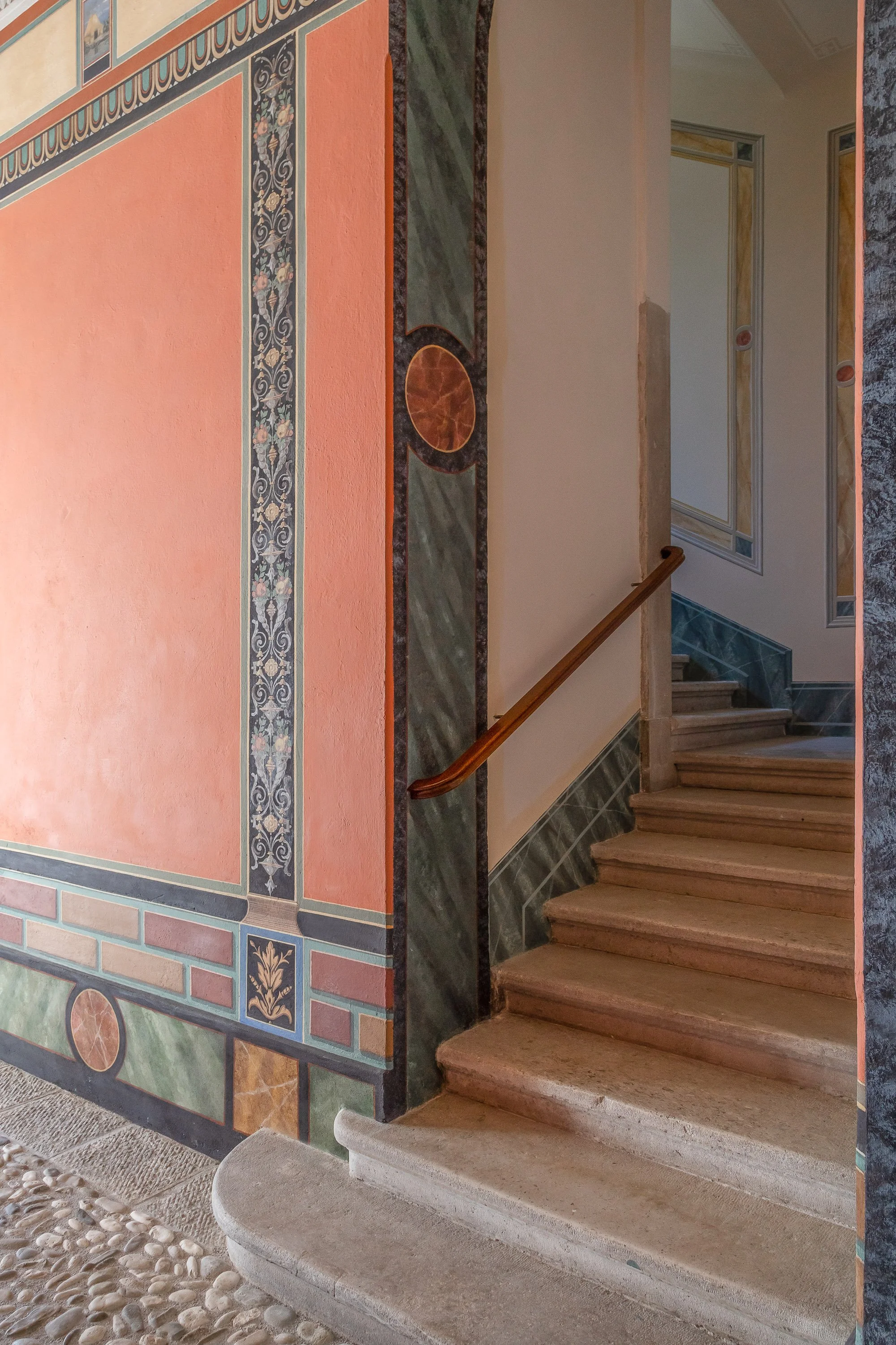 Scala in pietra con ringhiera di legno e pareti decorate con affreschi e mosaici.