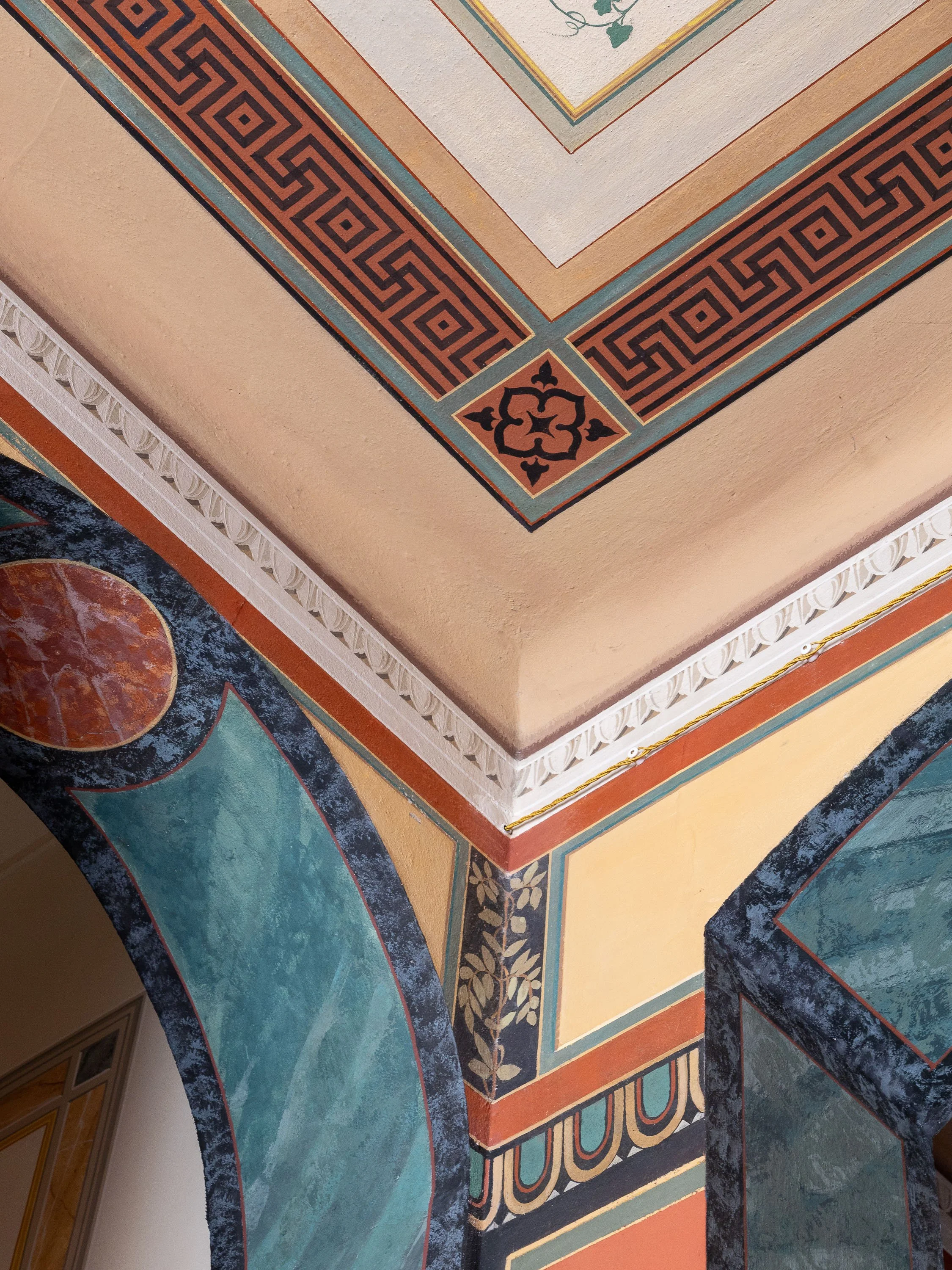 Resti di affreschi colorati con motivi geometrici e decorativi su soffitto e parete, con dettagli di cornici decorative e motivi floral e geometrici, in stile classico o antico.