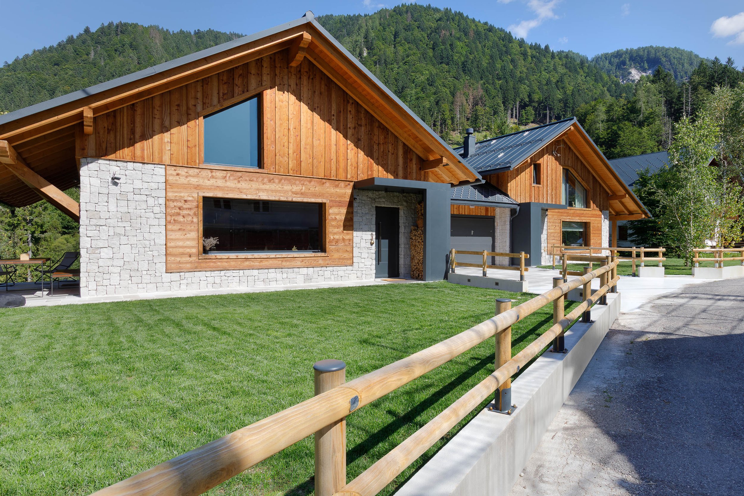 Casa in stile moderno con pareti in legno e pietra, tetto in metallo, grandi finestre e un cortile curato, circondata da montagne verdi.