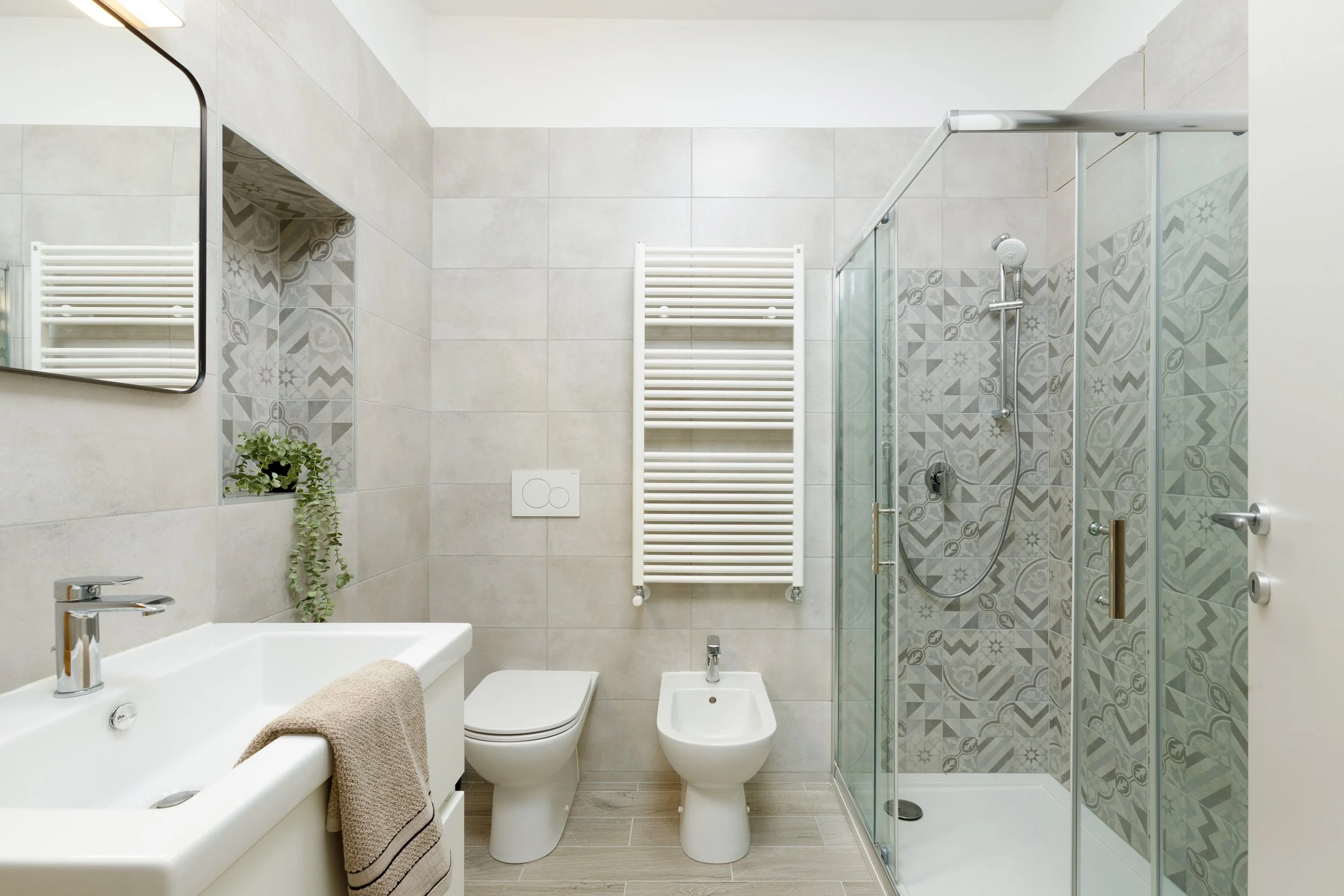 Bagno moderno con doccia in vetro, lavabo, asciugamano beige, specchio, WC, bidet e piastrelle decorative.