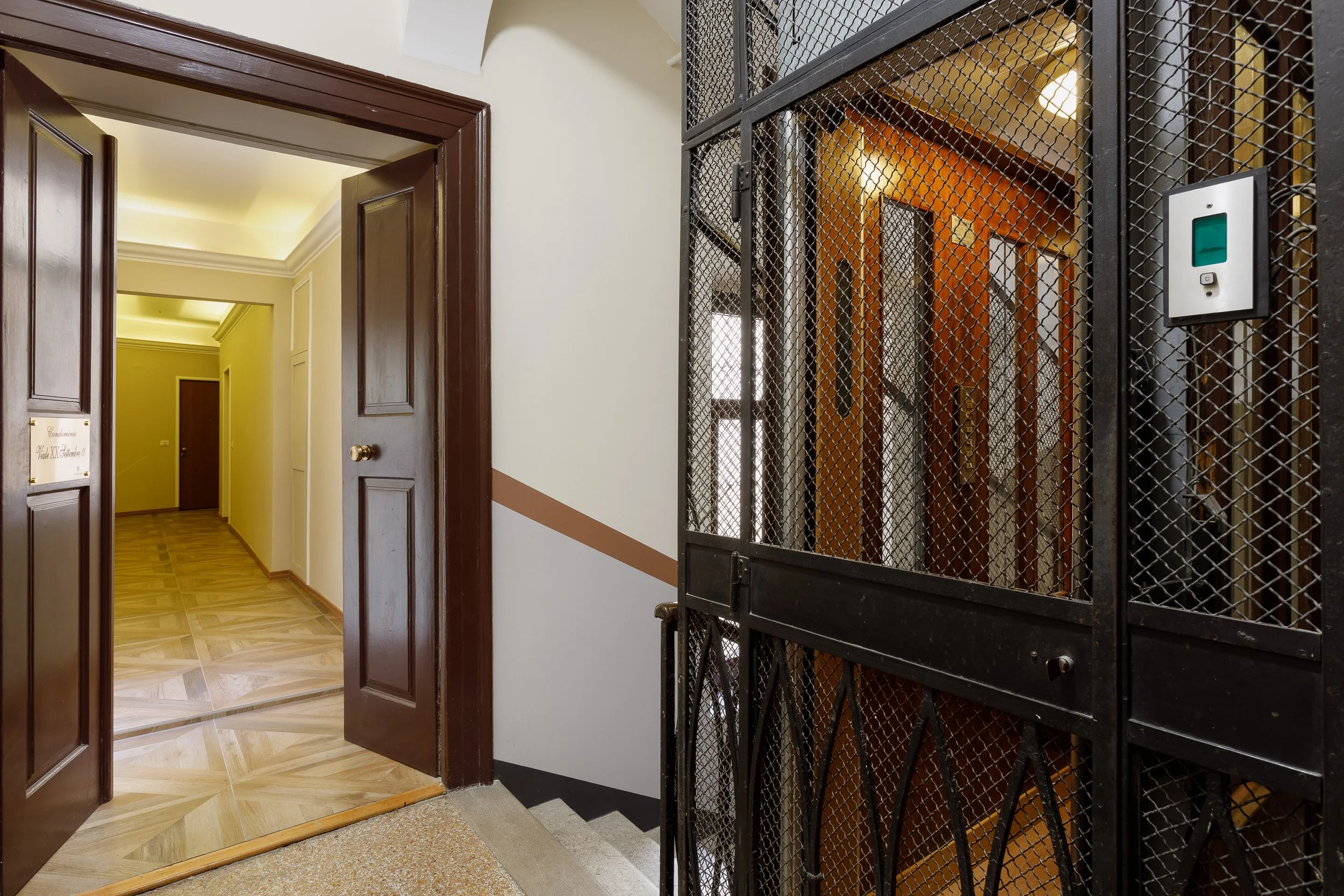 Corridoio di un edificio con ascensore storico a gabbia e porte in legno aperte.