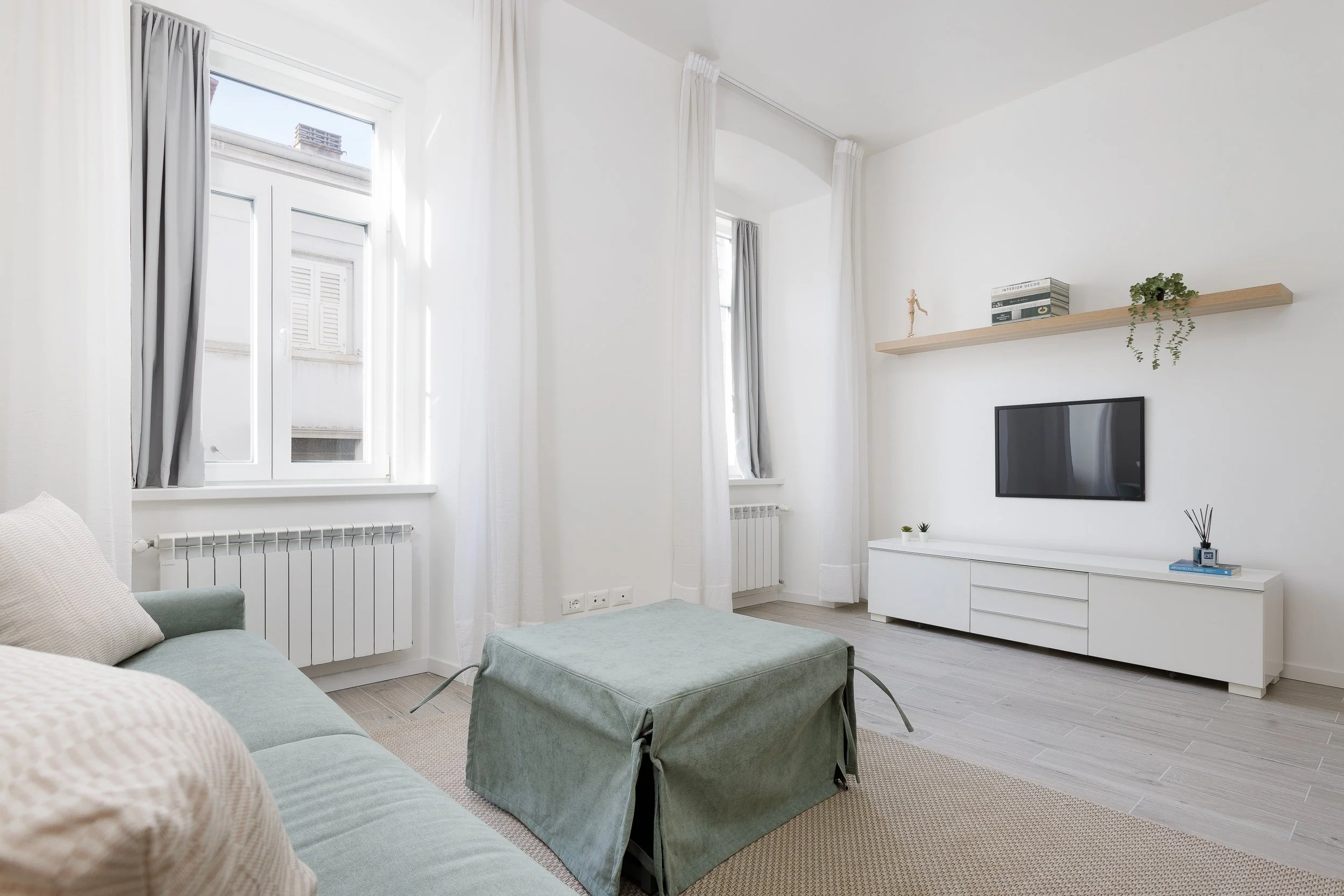 Soggiorno moderno con divano verde, finestra con tende leggere, tavolino imbottito, parete con mensola, TV montata, mobile bianco, decorazioni minimaliste e pavimento in legno chiaro.