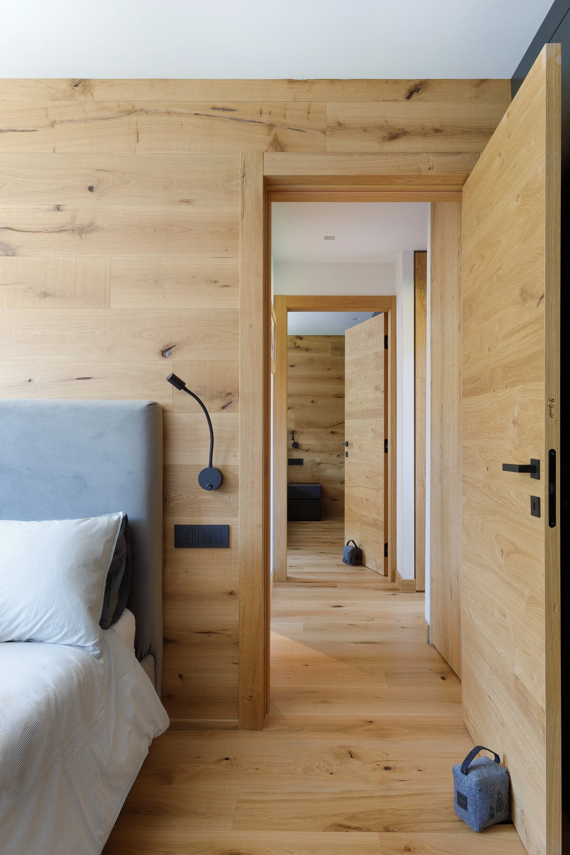 Camera con pareti e pavimenti in legno chiaro, letto con biancheria a righe, lampada da lettura nera, prese di corrente e interruttori visibili, porte in legno aperte che conducono a altre stanze.