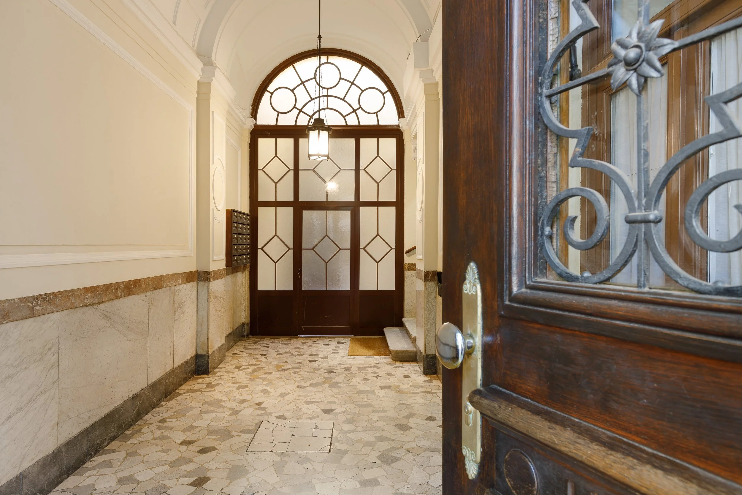 Ingresso di un edificio con porta in legno aperta, pavimento in marmo e pareti in marmo chiaro. Porta interna con vetrate e lampadario sospeso.