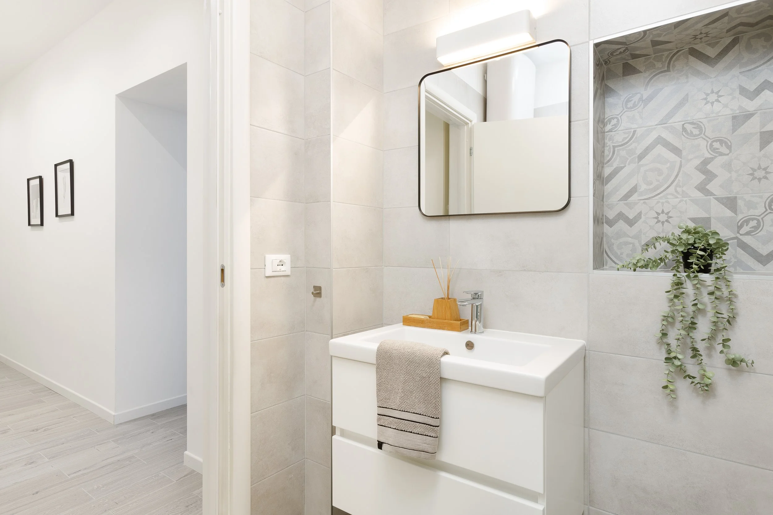 Bagno moderno con pareti piastrellate in grigio, specchio rettangolare, lavabo bianco con asciugamano, decorazione vegetale e accessori in legno.