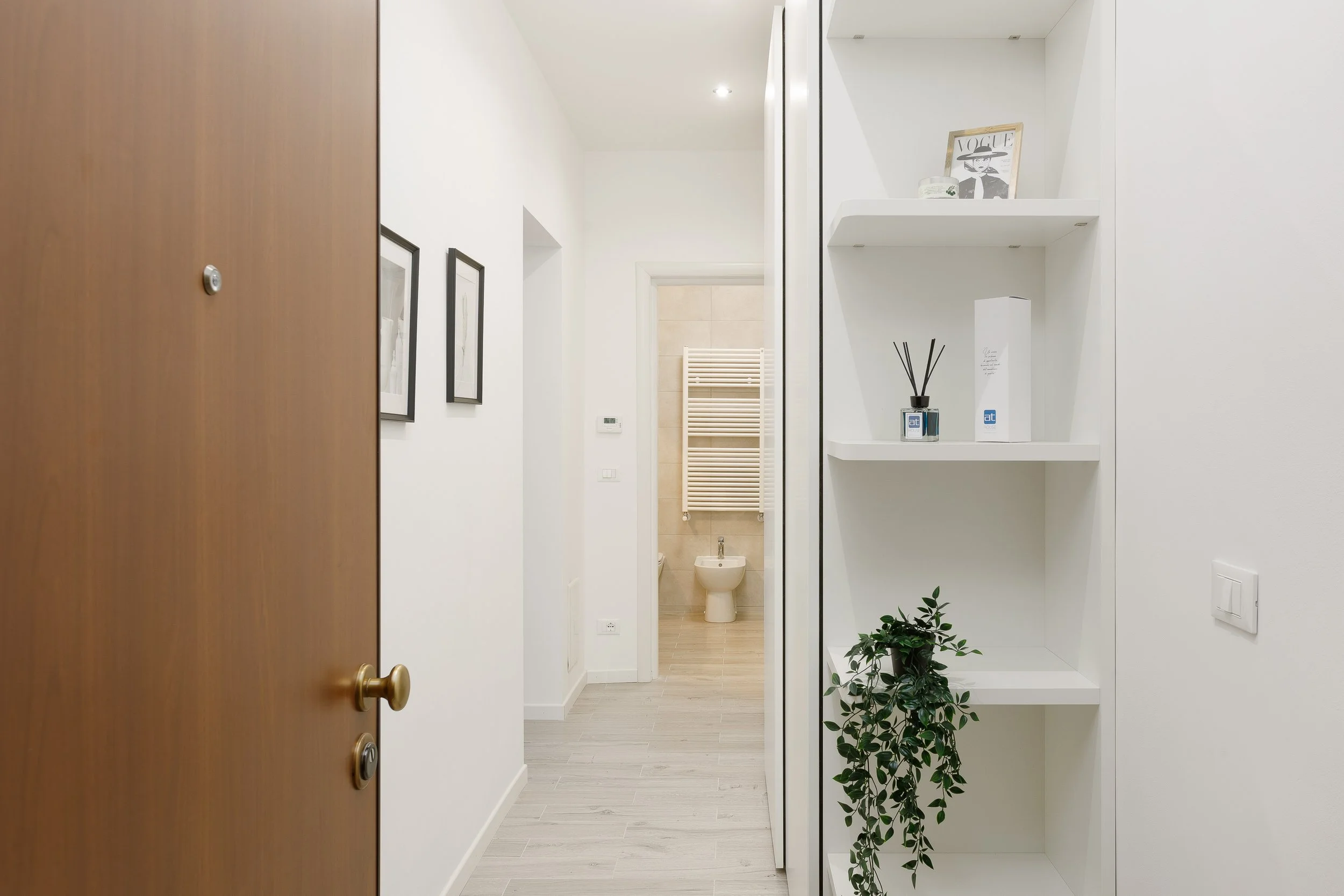 Ingresso di un appartamento con porta di legno, corridoio, scaffali bianchi con decorazioni, foto incorniciate alle pareti e vista di un bagno con sanitari e termoarredo.