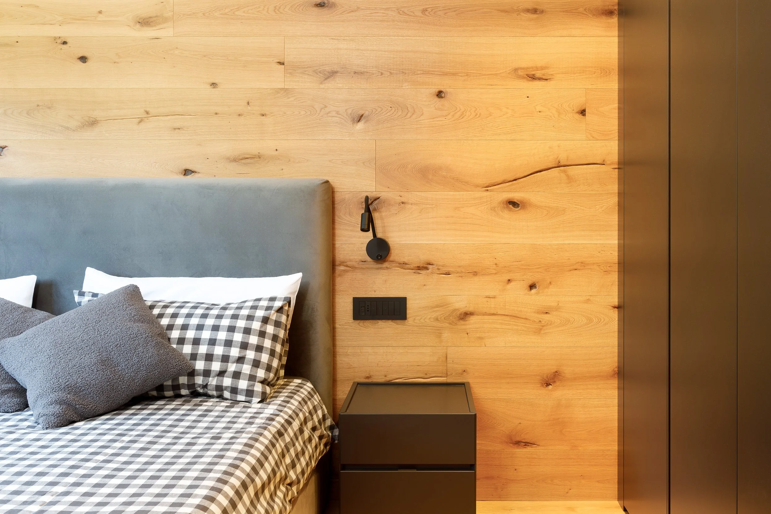 Interno di una stanza con parete di legno, letto con cuscini grigi e coperta a quadri, lampada da parete nera e una piccola parete attrezzata nera.