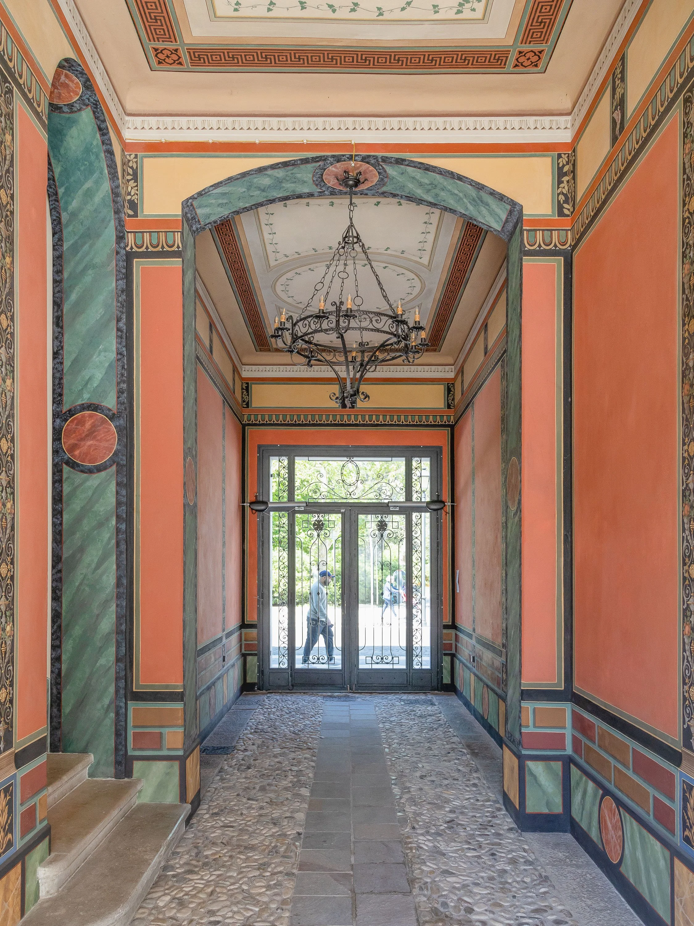 Ingresso decorato con pareti dipinte di colori caldi e soffitto con affreschi. C'è un lampadario in ferro battuto e una porta in vetro con inferriate.
