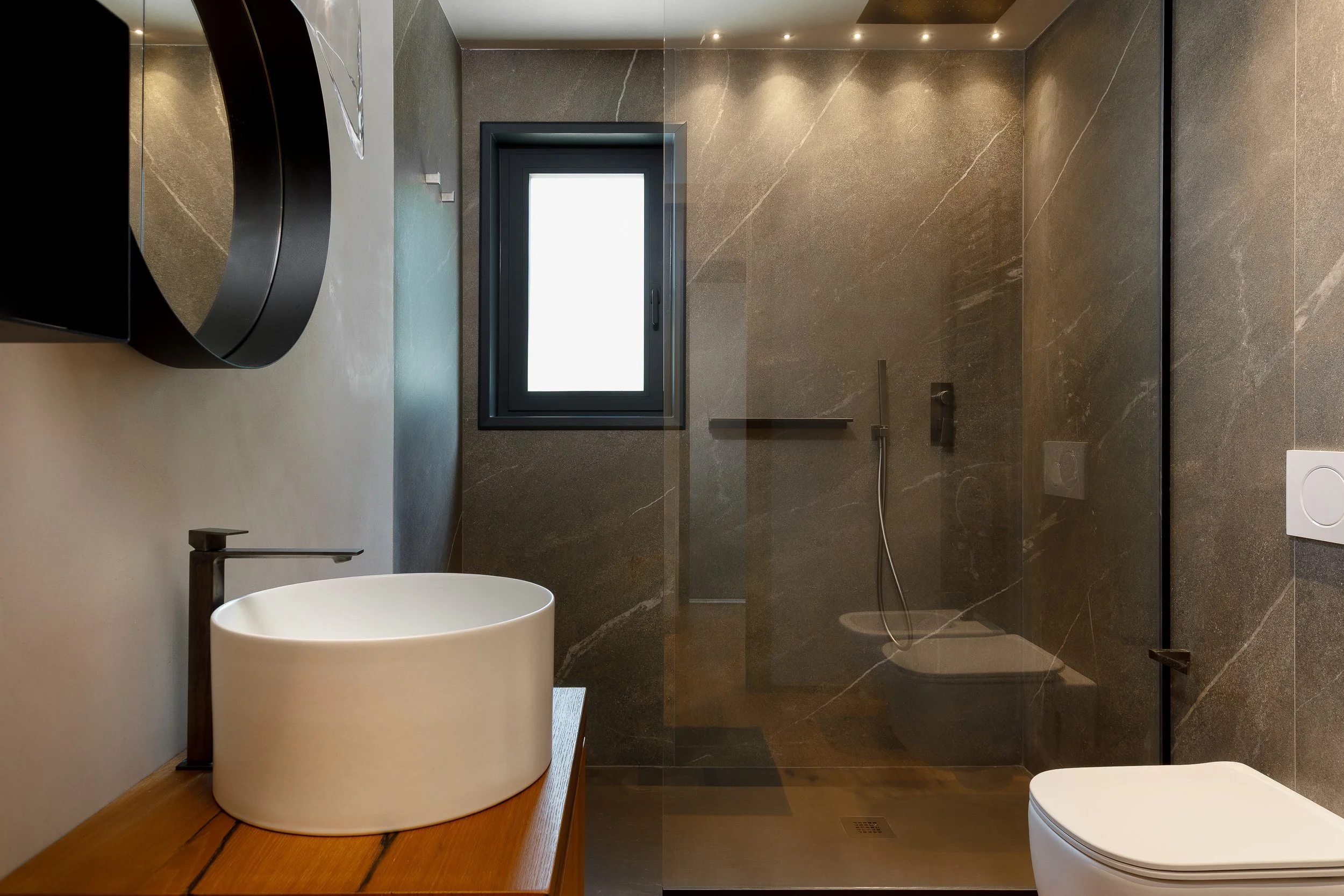 Interno di un bagno moderno con pareti in pietra scura, finestra, lavabo rotondo bianco su superficie di legno, WC e doccia con pareti di vetro e soffitto con luci LED.