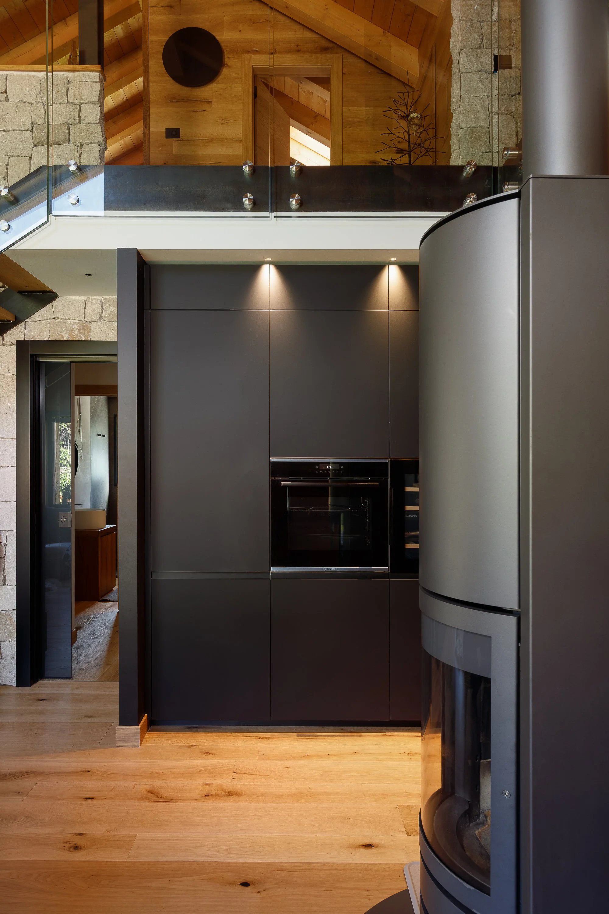 Interno di una casa moderna con pareti in pietra e legno, cucina con armadi grigi e dettagli in acciaio, pavimento in legno chiaro, soffitto con travi in legno e una scala che porta al piano superiore.
