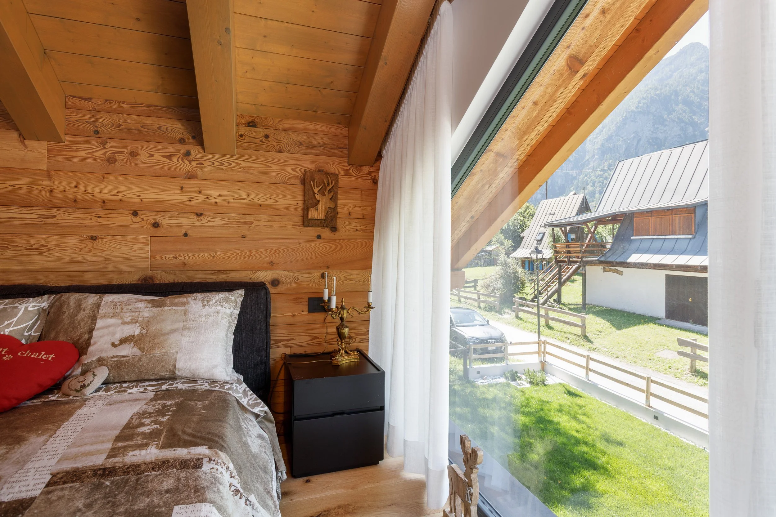 Interno di una camera con pareti di legno, letto con cuscino rosso, lampada dorata e finestra grande con vista sulle case di montagna e il verde del prato.