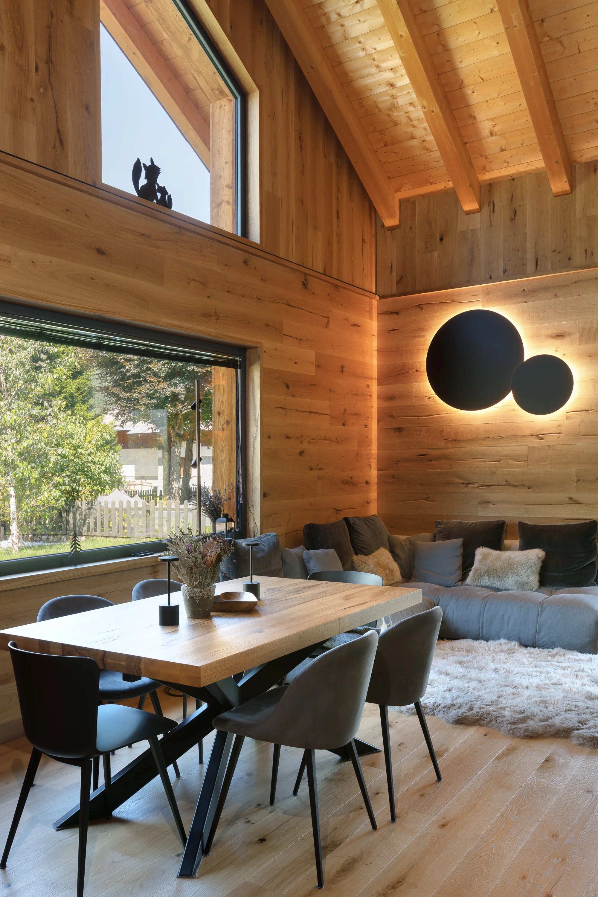 Interno di un soggiorno con pareti e soffitto in legno, grandi finestre che mostrano un giardino con alberi, un tavolo in legno con sedie nere, un divano grigio con cuscini e una lampada a soffitto con due sfere nere che emettono luce. Decorazioni co