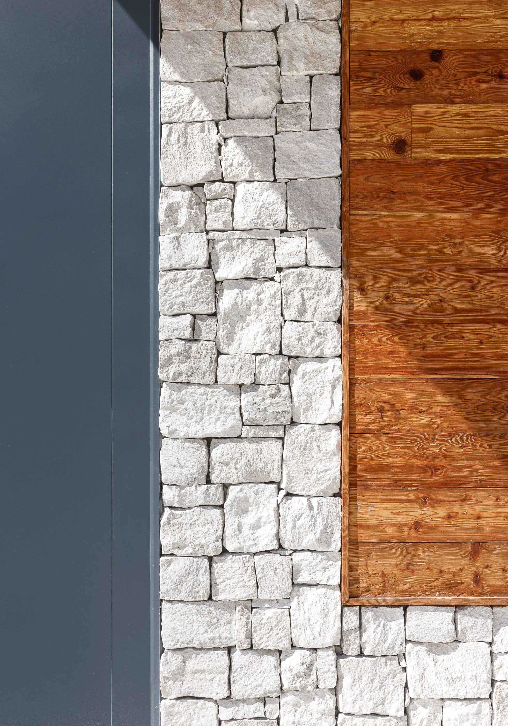 Un muro decorativo con tre sezioni: una sezione di pannelli di metallo grigi, una sezione di pietre bianche disposte in modo irregolare e una sezione di pannelli di legno di colore marrone con venature visibili.