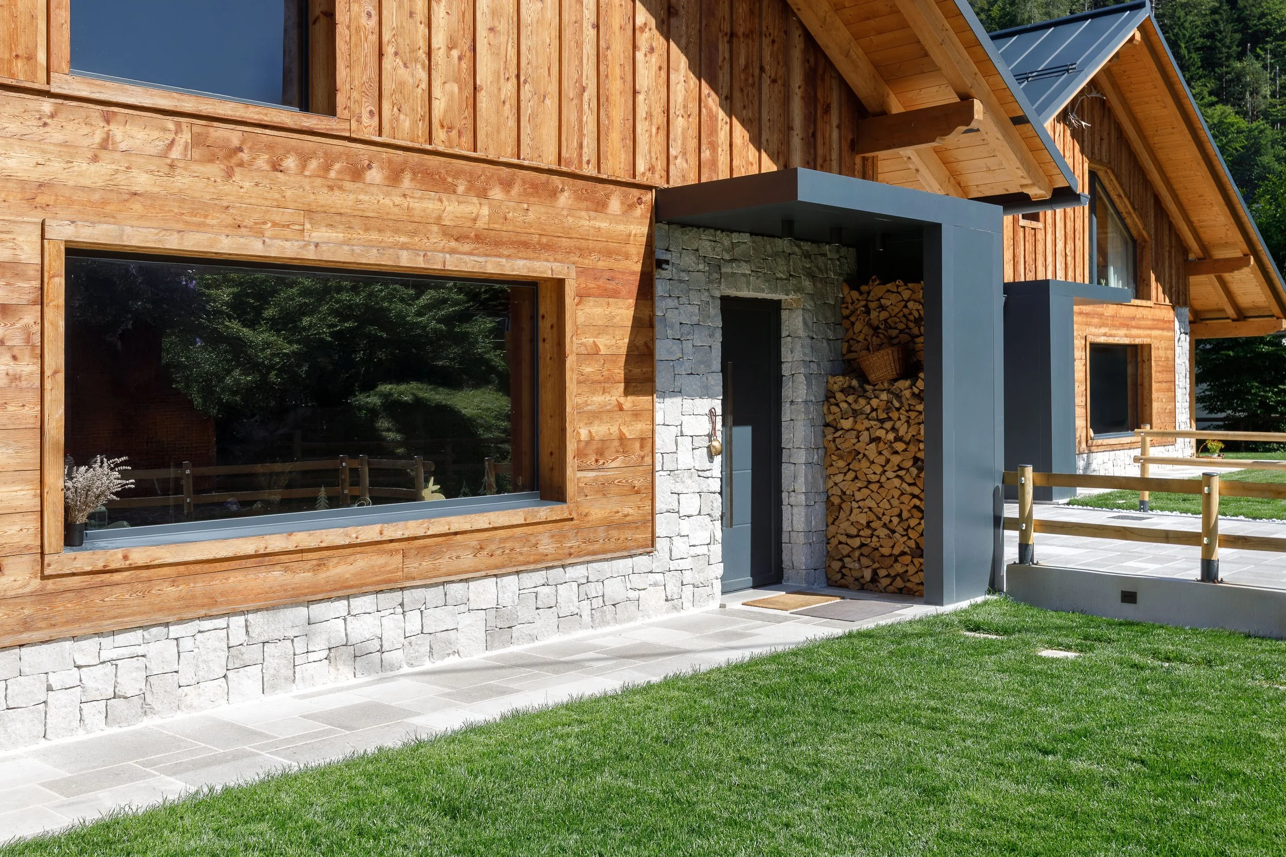 Casa in stile alpino con pareti in legno e pietra, finestra grande e pila di legna accanto alla porta d'ingresso, con un cortile e prato verde circostante.