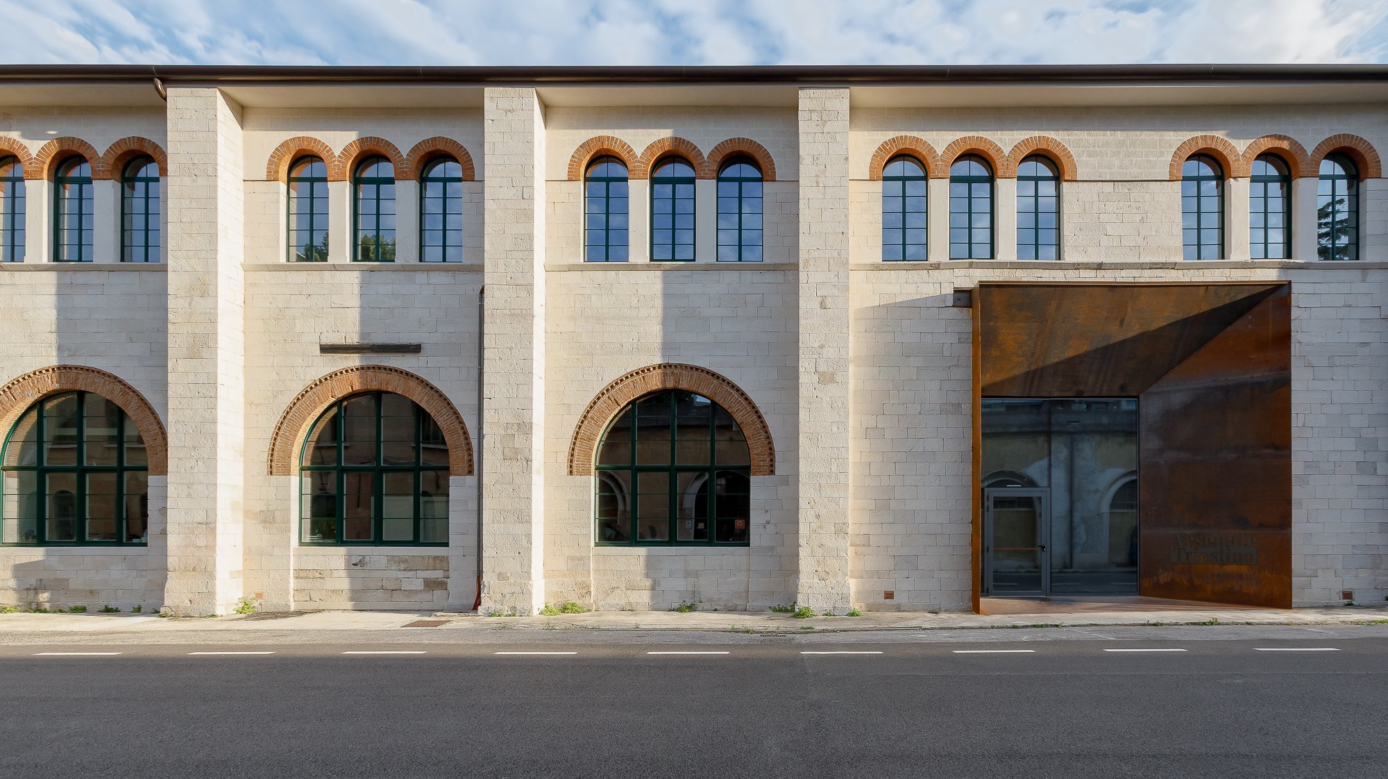 Edificio con pareti in pietra e finestre ad arcata con cornici in mattoni, ingresso con porta di vetro e struttura in ruggine, strada di fronte.