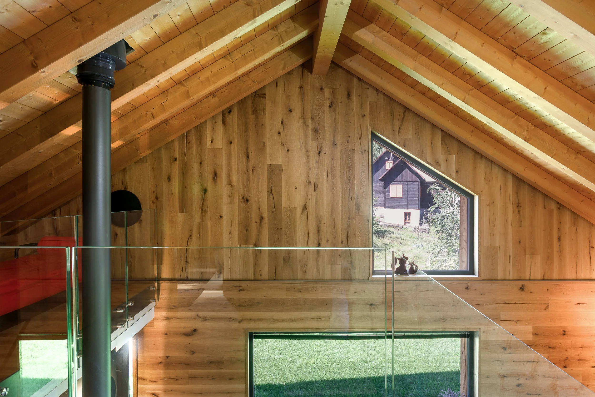 Interno di una casa di legno con pareti e soffitti in legno chiaro, finestra grande che mostra un giardino verde e un edificio in legno sullo sfondo.