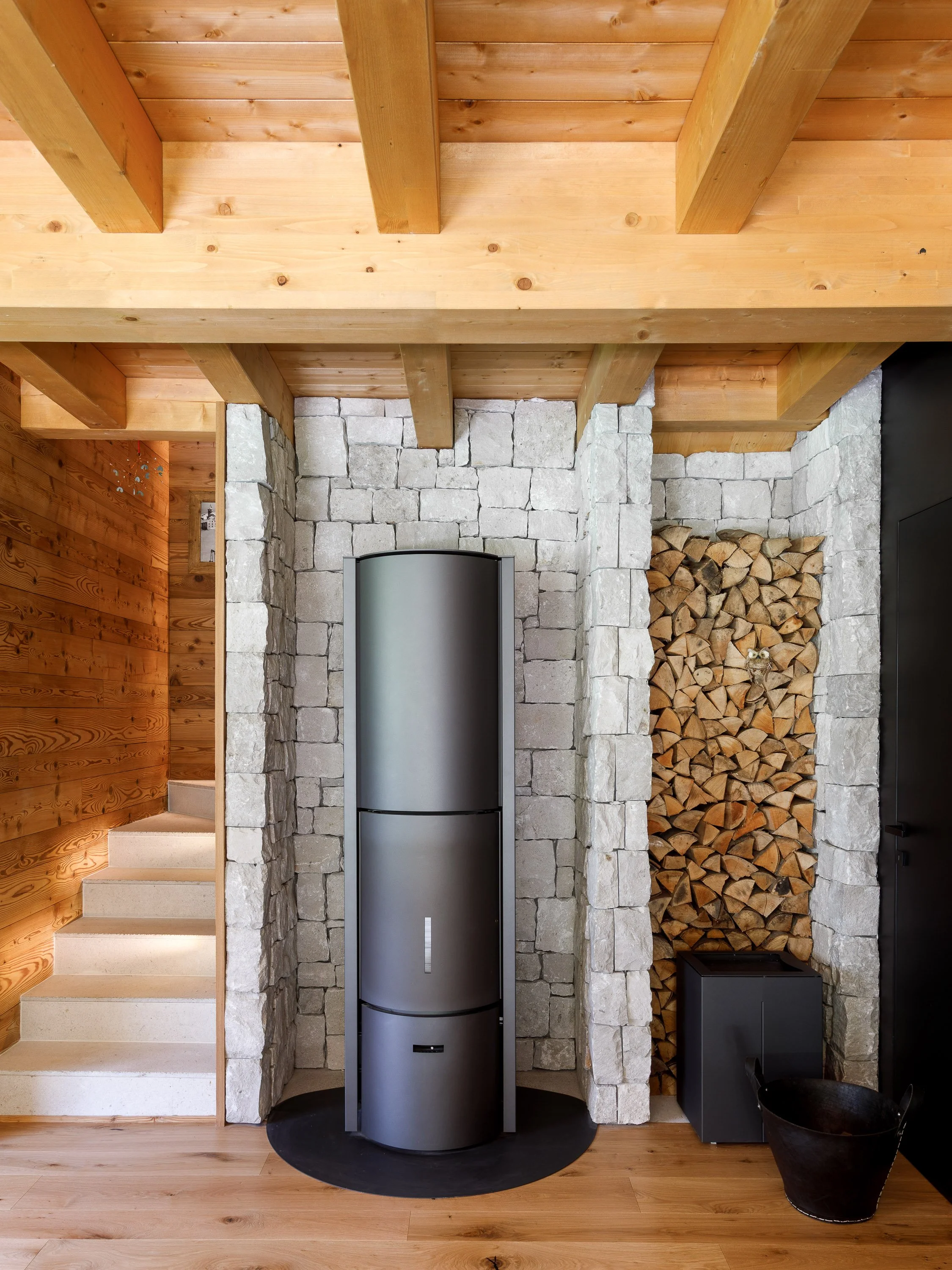 Interno di una casa con pareti in pietra, camino in metallo, storico di legno e un muro di legno con legna accatastata, pavimento in legno.