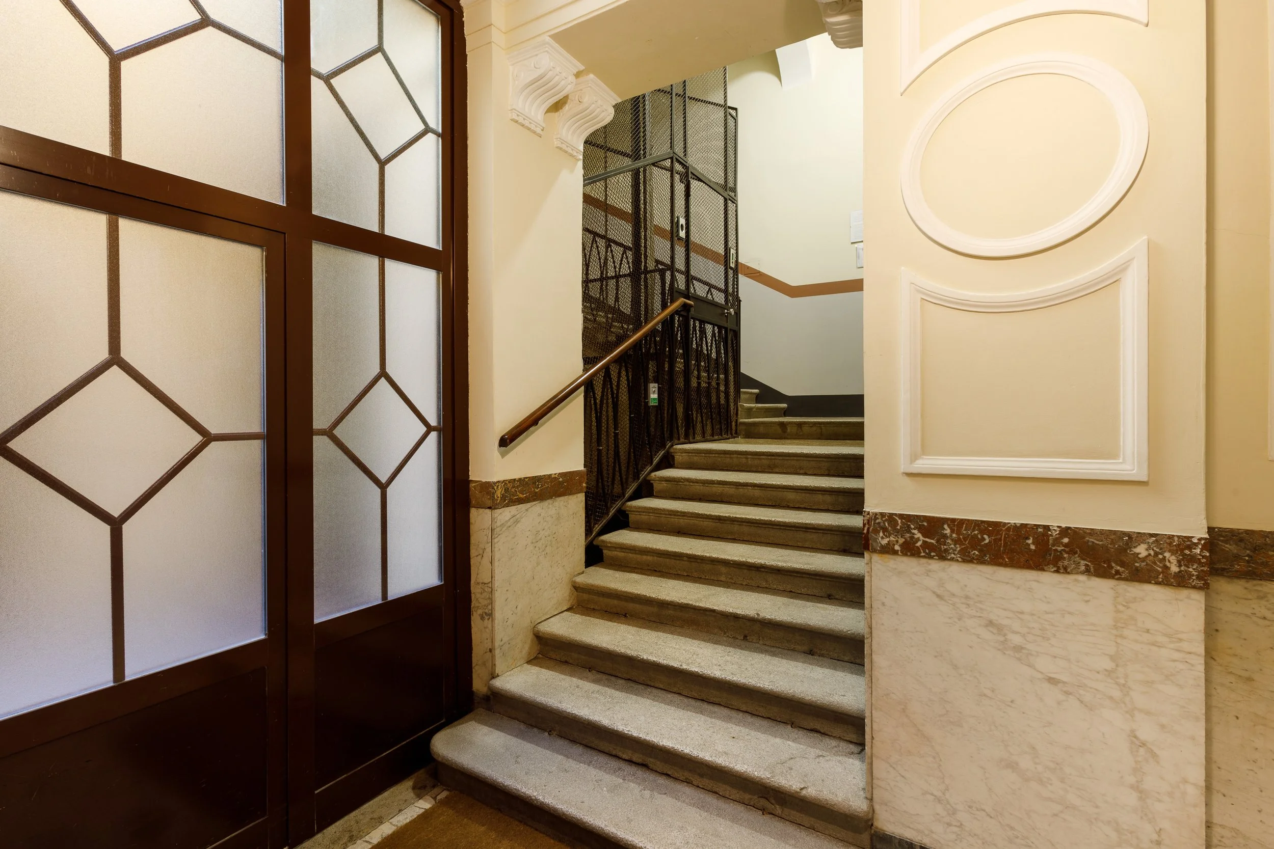 Ingresso di un edificio con scala in marmo, corrimano in metallo e pannelli di vetro decorati.