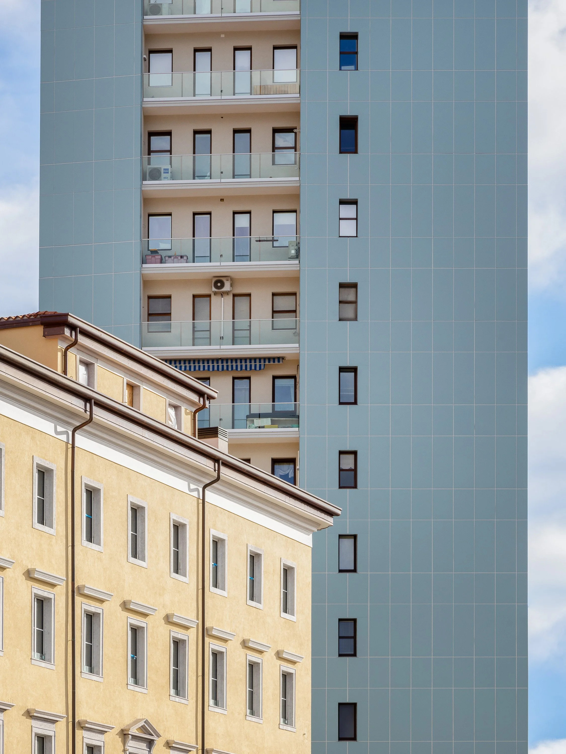 Due edifici residenziali affiancati, uno color oro chiaro con finestre rettangolari e decorazioni, l'altro moderno e alto con facciata di vetro e balconi, alcuni con mobili e aria condizionata.