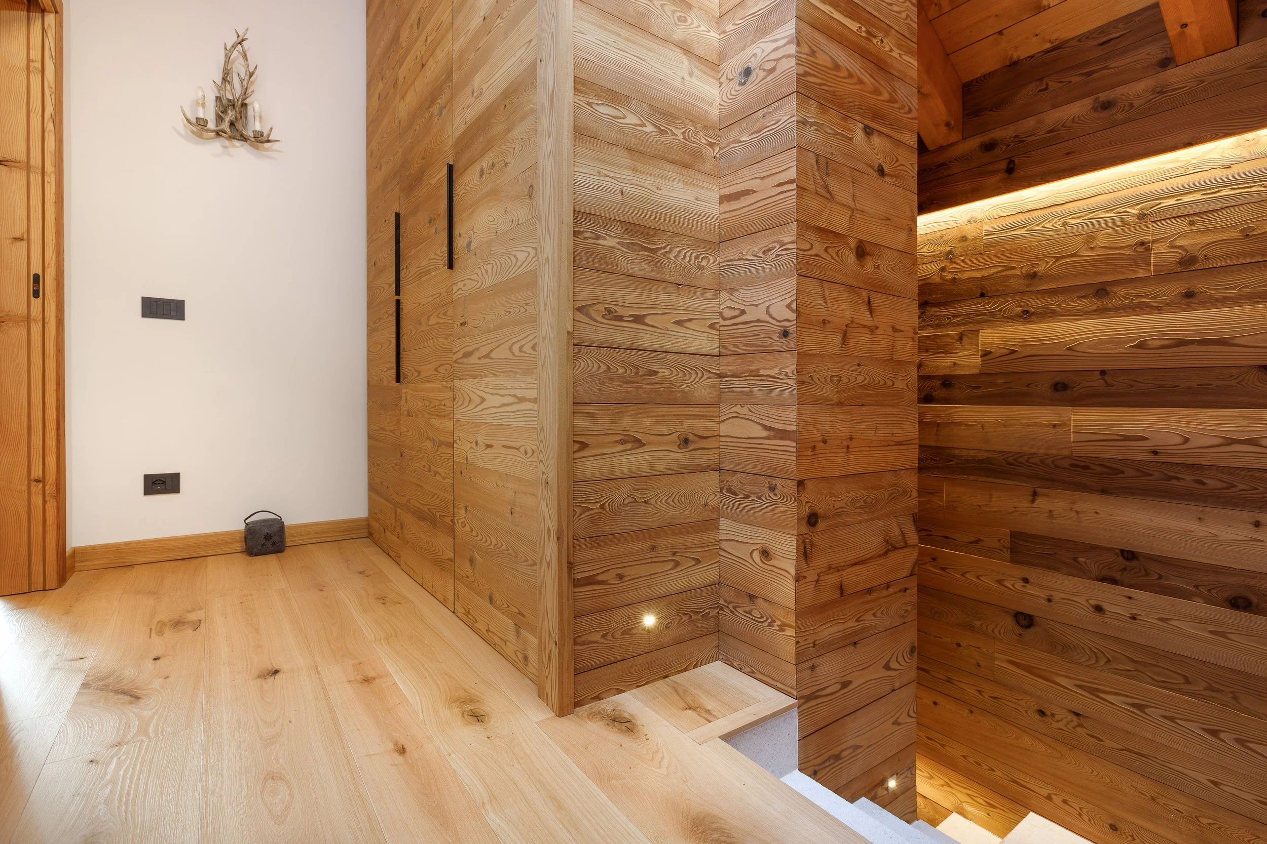 Interno di una casa con pareti e soffitto rivestiti di legno, con un'illuminazione nascosta e un pavimento in legno chiaro.
