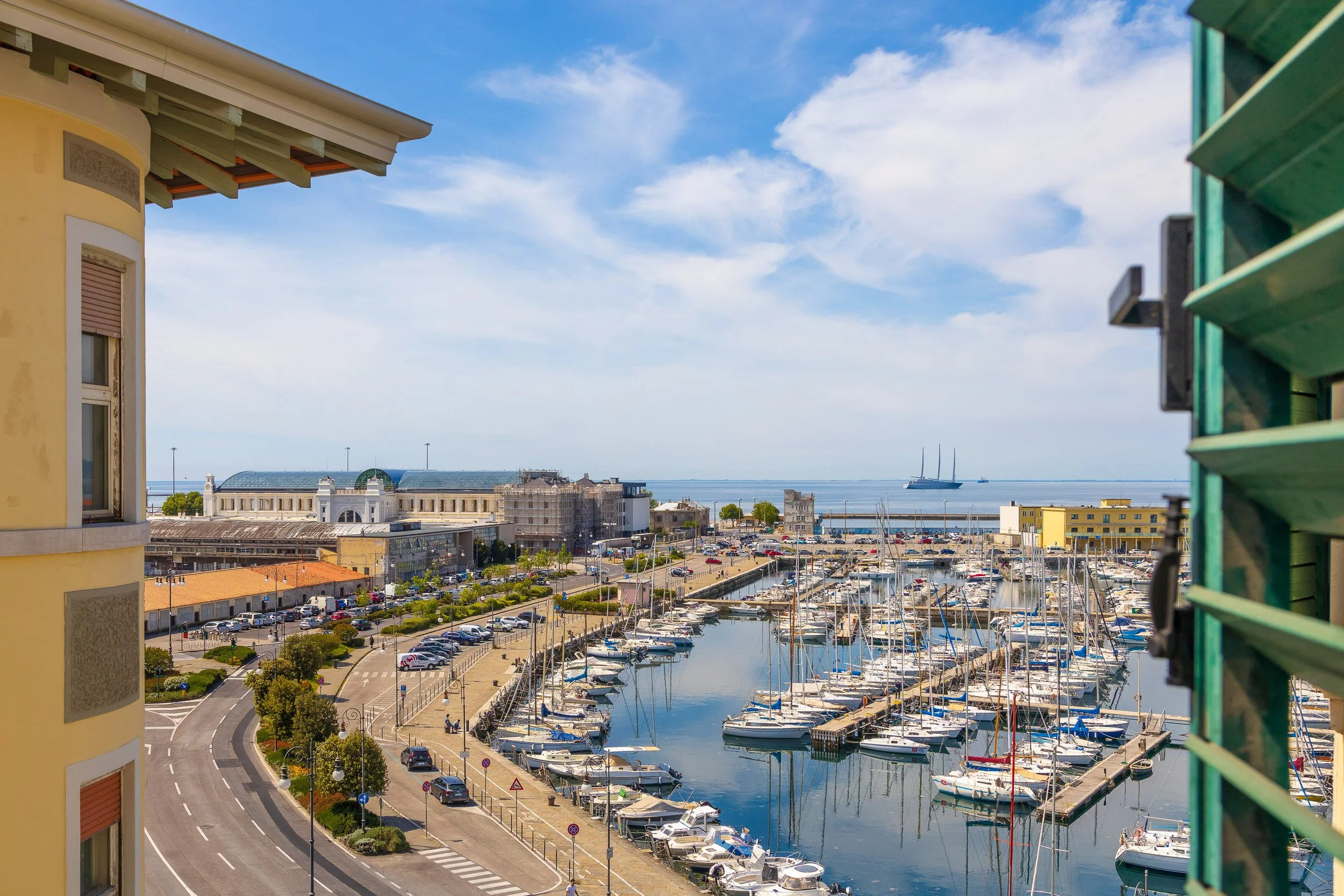 Veduta di un porto con numerose barche ormeggiate, un edificio storico, una strada trafficata e il mare sullo sfondo con una nave a vela.
