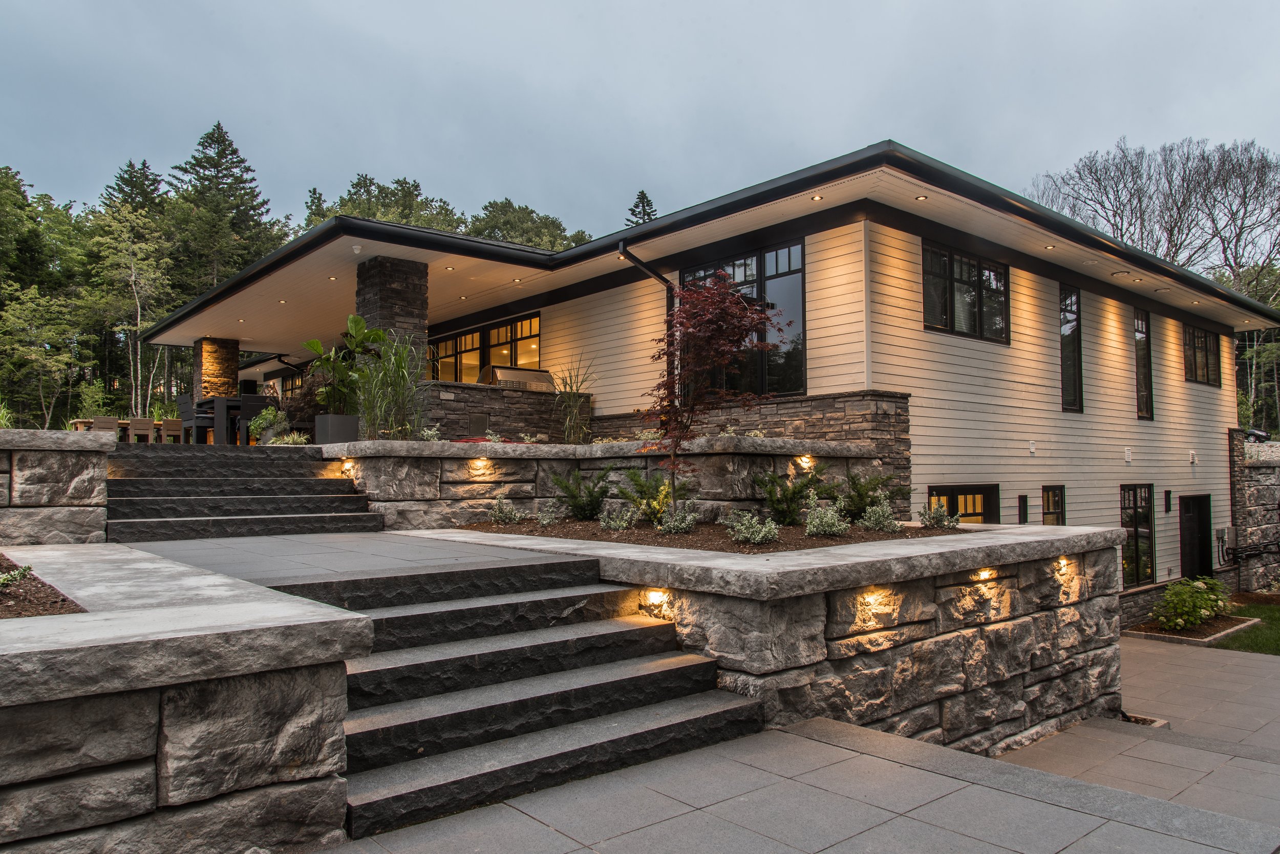 Modern bungalow house with wood exterior, covered deck, doors, black-framed windows, and a porch with a glass railing, surrounded by trees.