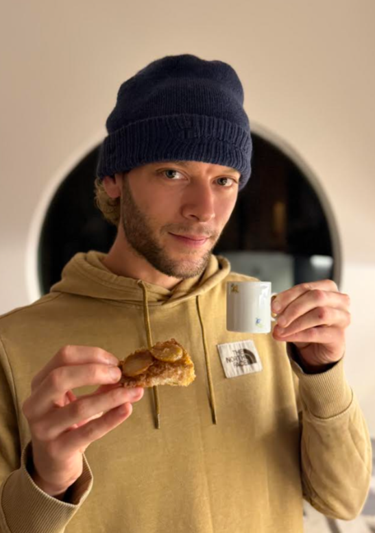 A young man wearing a navy beanie and beige hoodie holding a small white coffee cup in one hand and a piece of toast with jelly in the other, standing indoors with a circular window in the background.