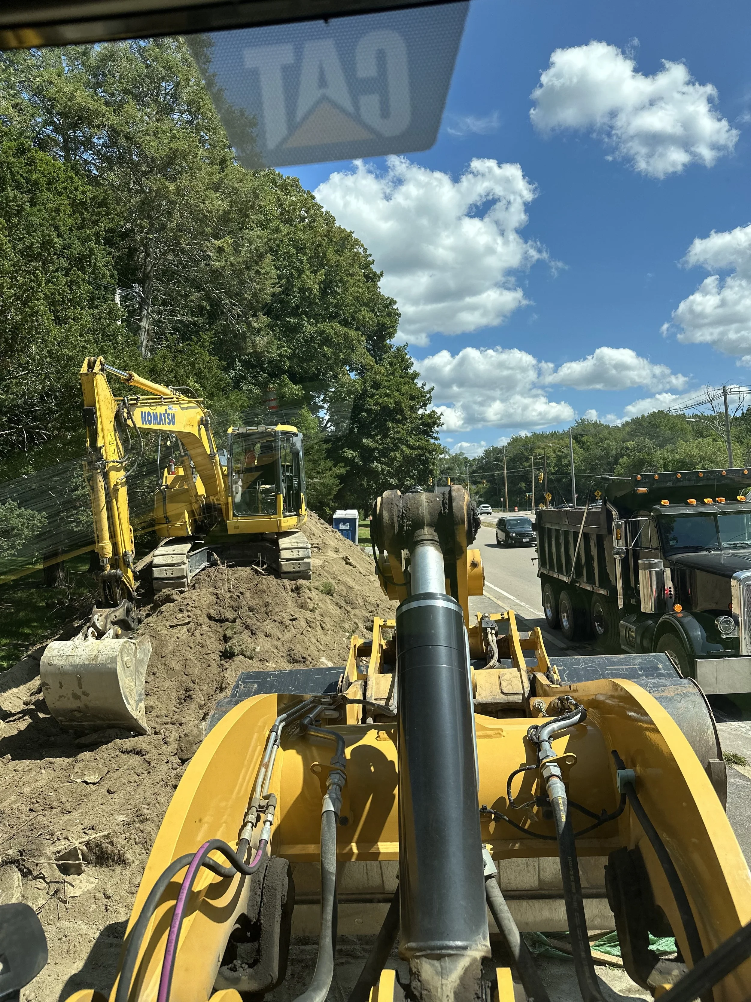Construction site showing a yellow Komatsu excavator digging into the ground, with a large rock, and a black dump truck nearby on a road with trees and cloudy sky in the background.