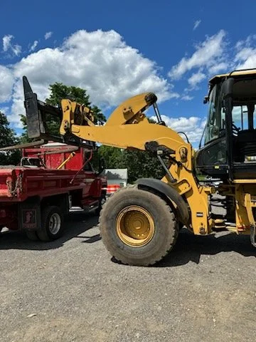 Construction vehicle and truck on a gravel surface under a partly cloudy sky.